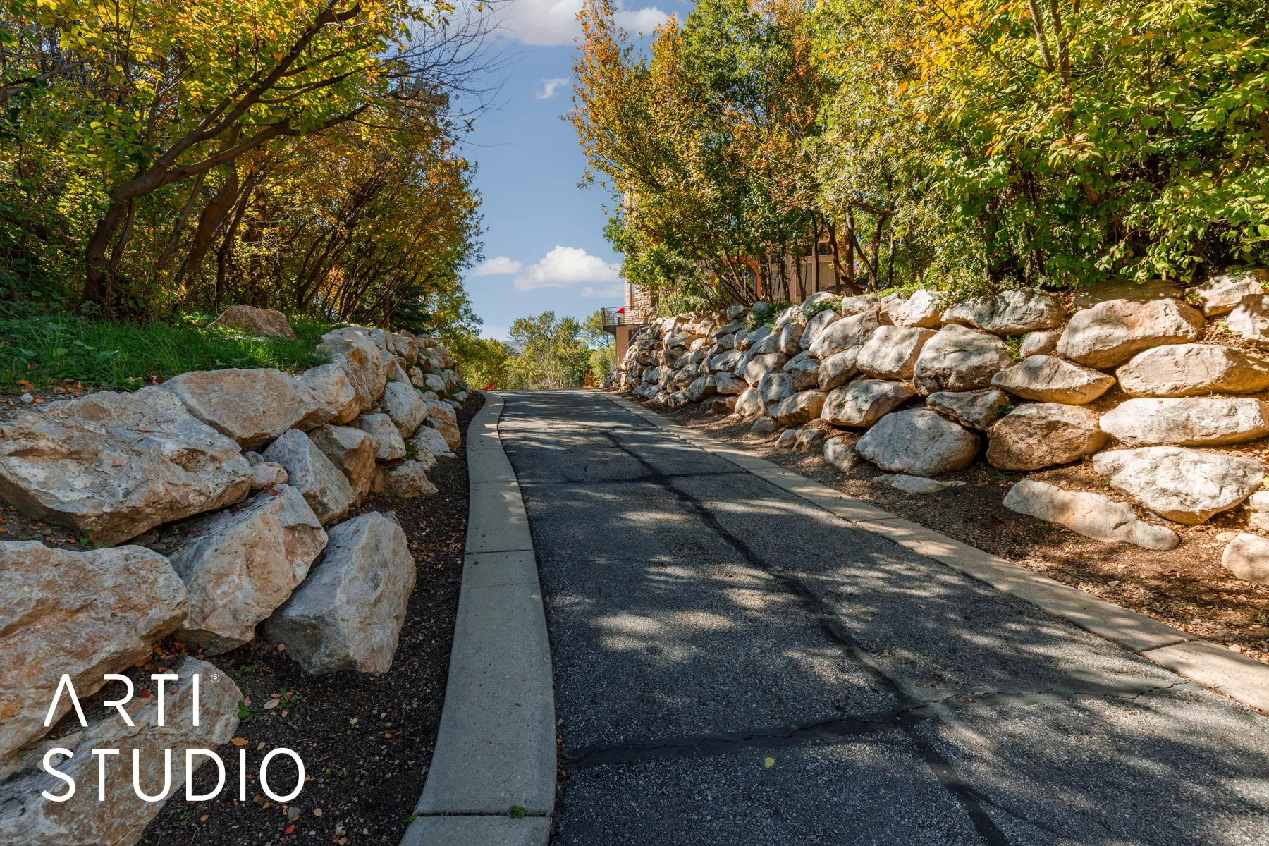 Asphalt driveway featuring concrete curbing, stone retaining walls, and mature deciduous trees