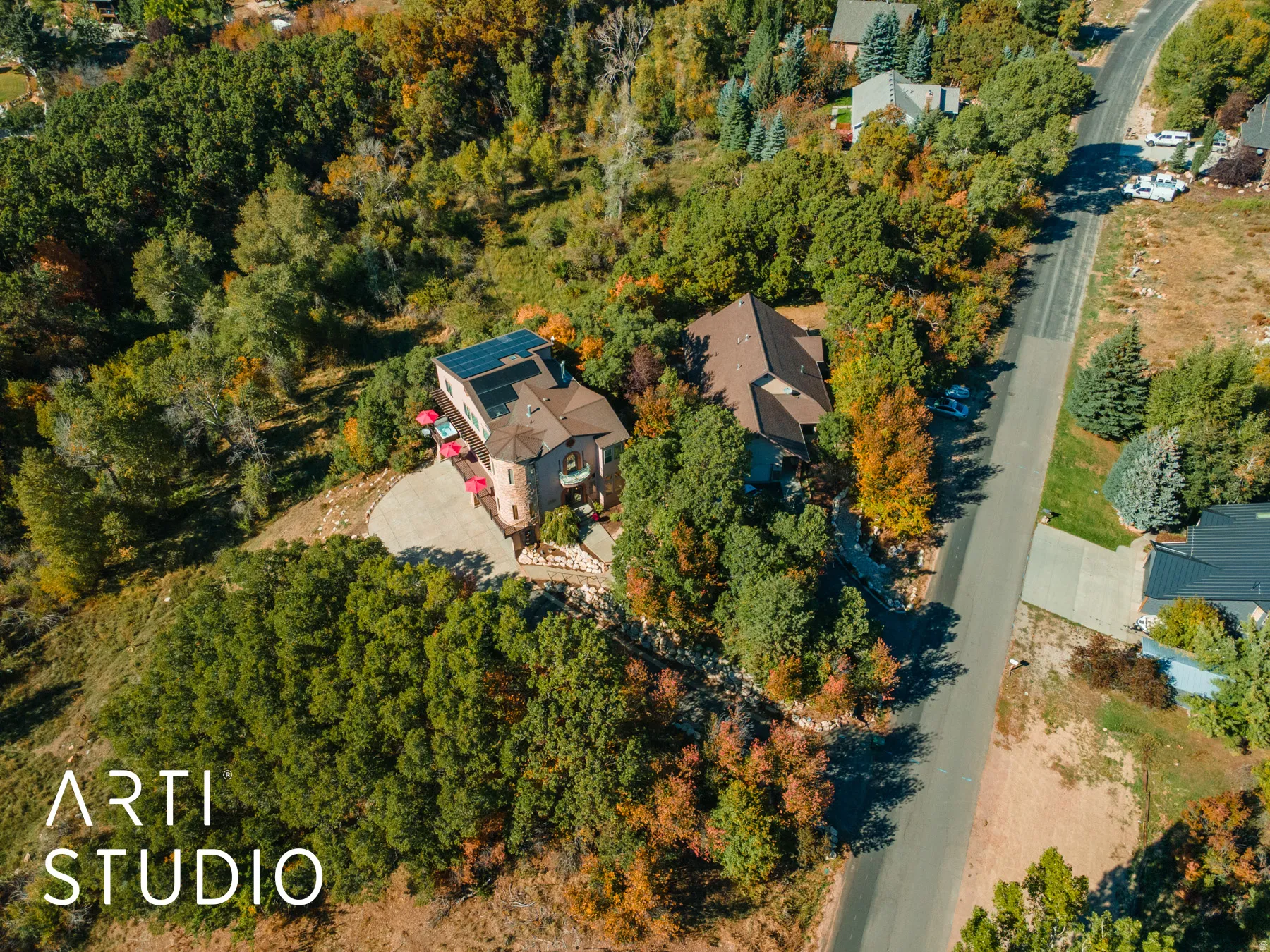 Hillside property featuring a stone turret exterior, a multi-paneled roof-mounted solar array, a curved paved driveway, and mature surrounding trees