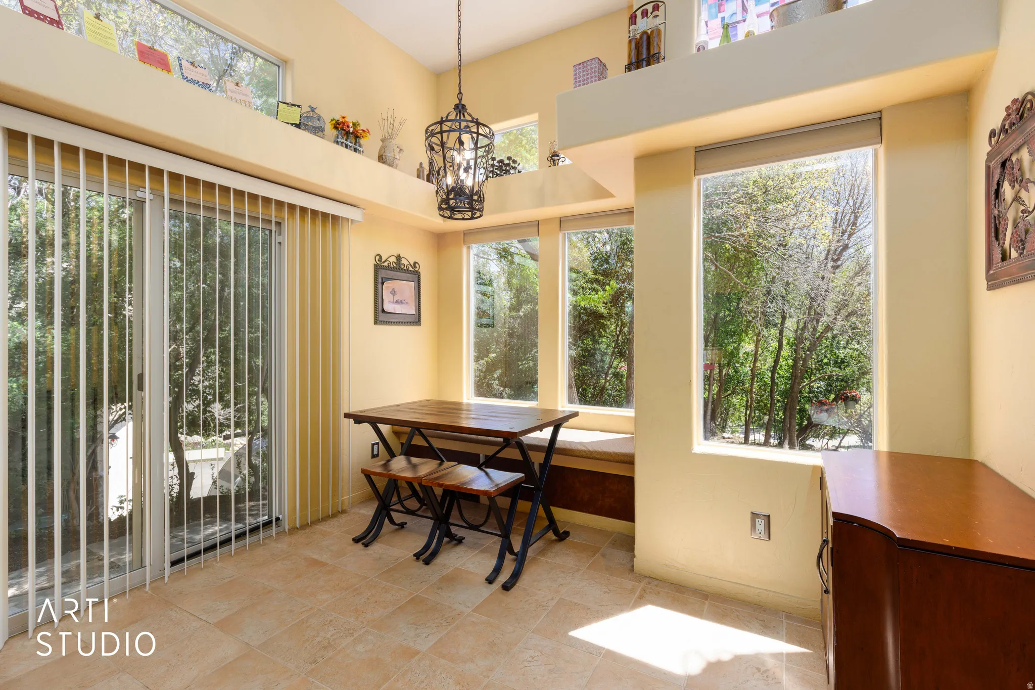 Sunlit interior space featuring floor-to-ceiling windows, a built-in window seat, a sliding glass door with vertical blinds, a vaulted ceiling, and tile flooring