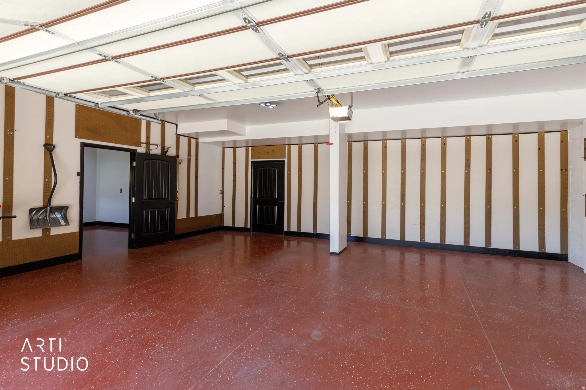 Spacious garage featuring a red speckled epoxy floor, white walls with wood-panel accents, two dark paneled doors, and exposed ceiling beams