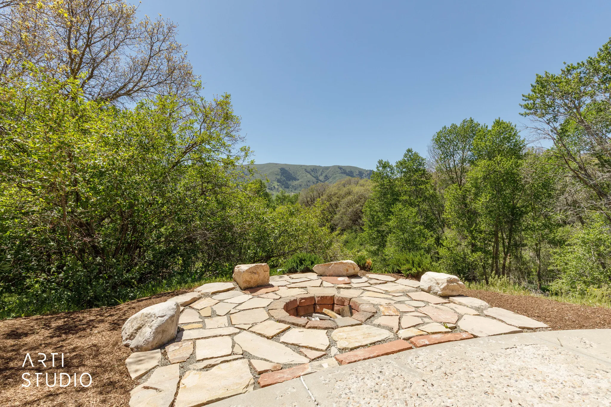 Outdoor fire pit area featuring a natural stone patio and brick fire ring, surrounded by lush trees and distant mountains