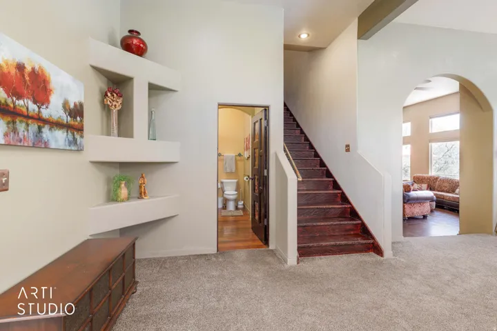 Carpeted entry area featuring a rich wood-finish staircase, built-in shelving, an arched doorway, and a visible powder room with wood-finish flooring