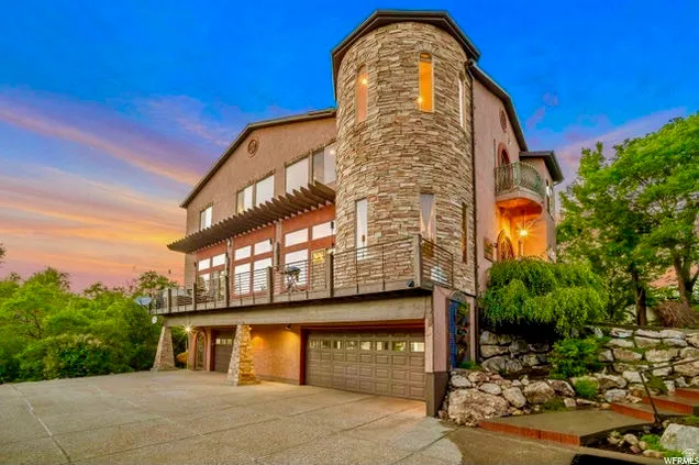Stucco exterior featuring a stone-clad turret, multiple balconies with metal railings, a pergola-covered patio, and an integrated multi-car garage