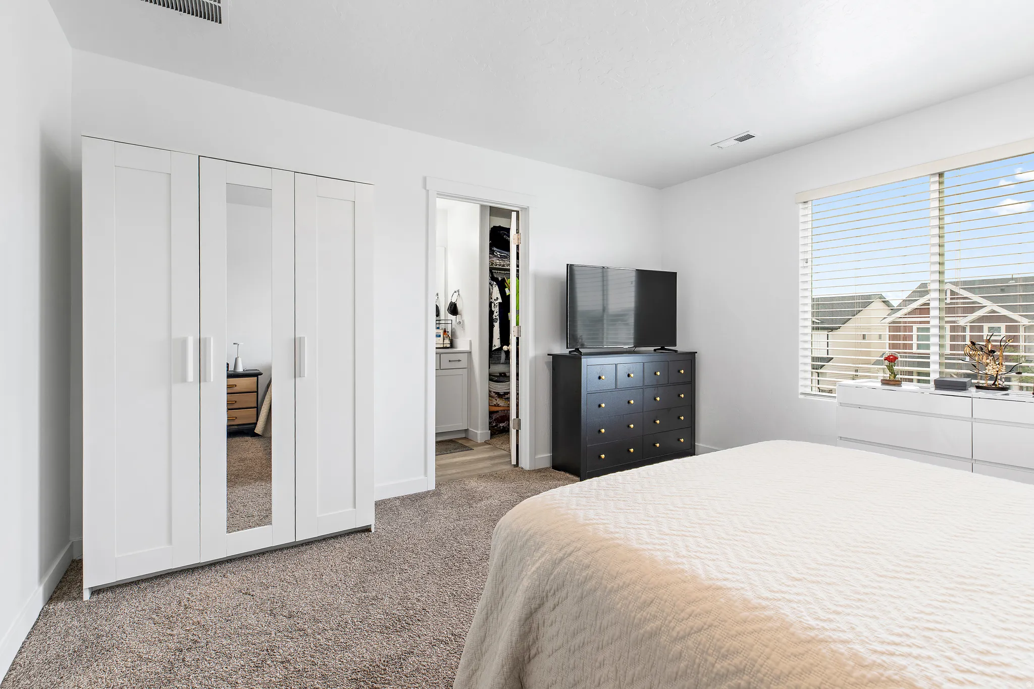 Carpeted bedroom with a large window and blinds, featuring white walls and a visible walk-in closet with built-in storage