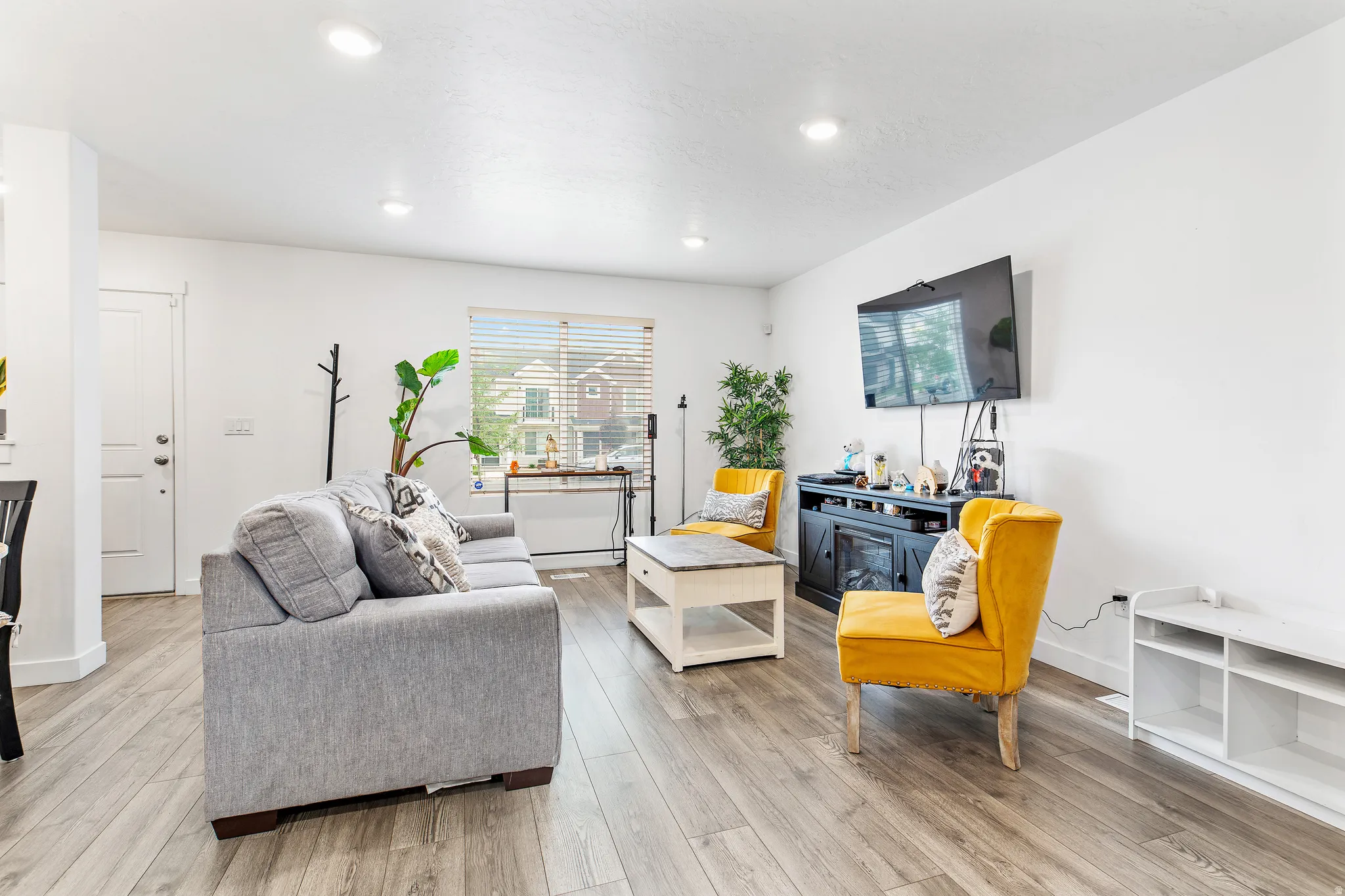 Open living area featuring wood-finish flooring, recessed ceiling lighting, a prominent window with blinds, and a white interior door