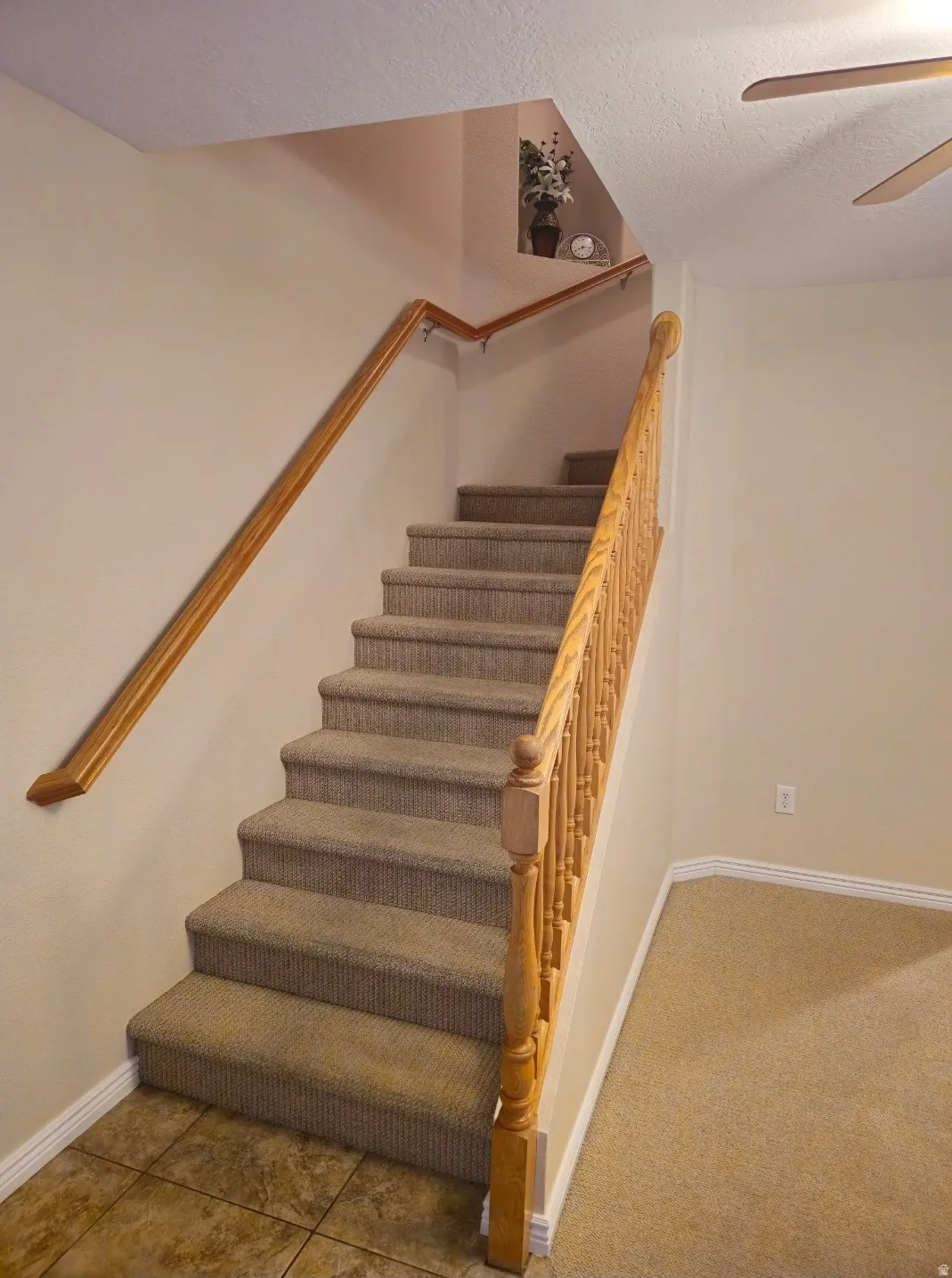 Carpeted staircase with light wood handrails and balusters, featuring a tiled landing at the base