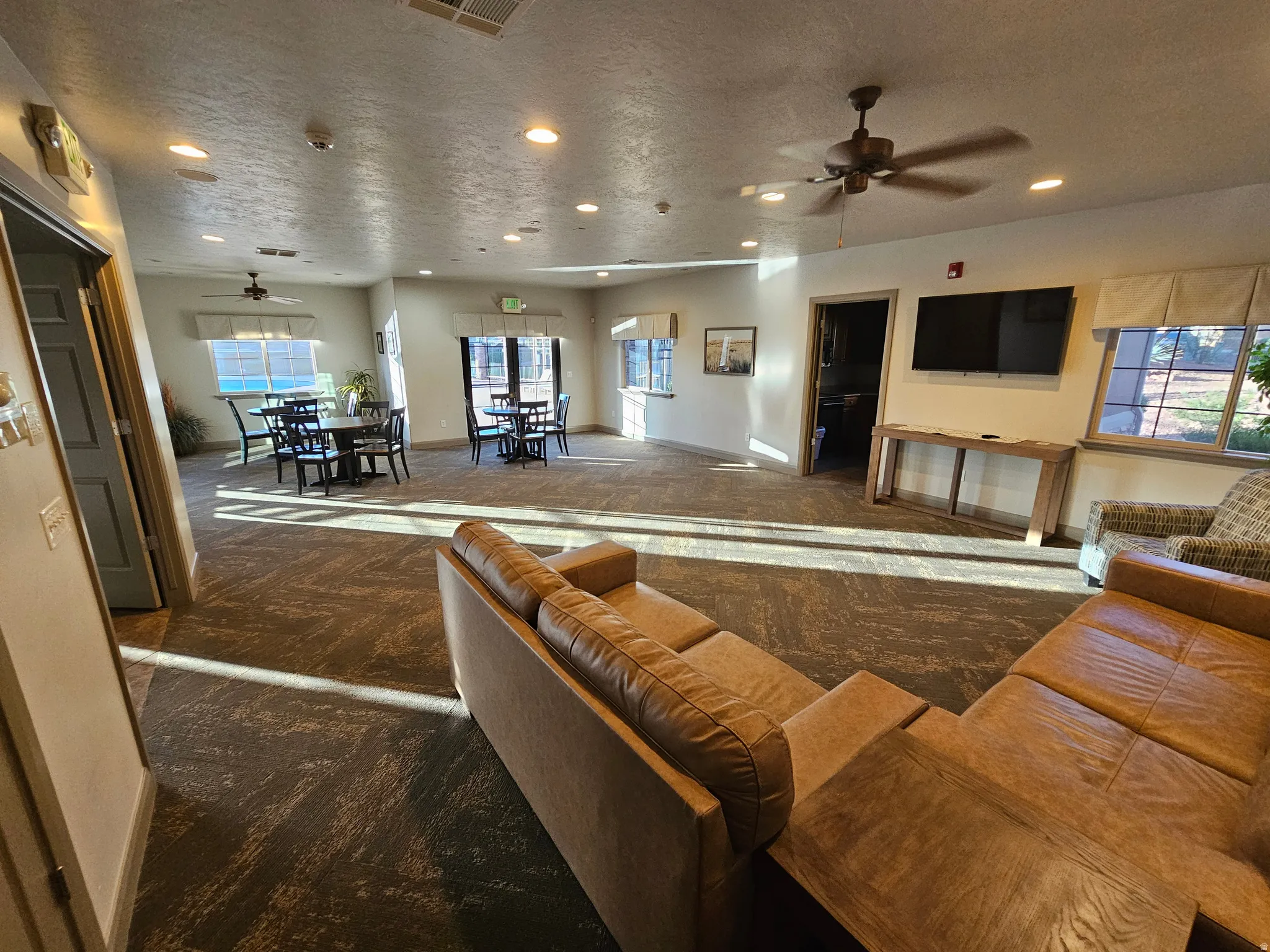 Spacious common area featuring textured carpeting, multiple ceiling fans, recessed lighting, and large windows with privacy blinds