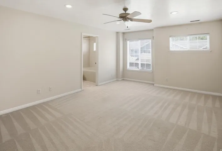 Spacious room featuring neutral carpeting, a ceiling fan with integrated lighting, two recessed ceiling lights, and multiple windows with horizontal blinds
