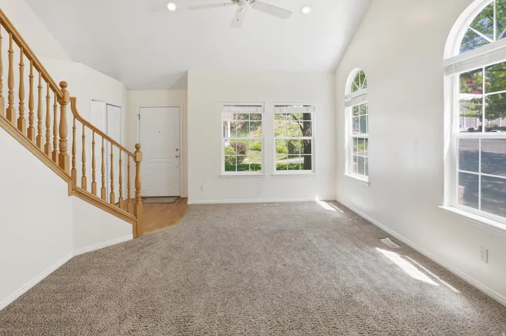 Spacious living area featuring vaulted ceilings, light-hued carpeting, multiple arched windows, a front entry door with wood-finish flooring, and a natural wood staircase with balusters