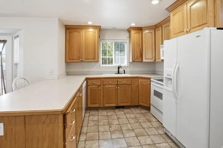 Kitchen featuring wood cabinetry, light-toned countertops, white appliances, recessed lighting, and a window above the sink