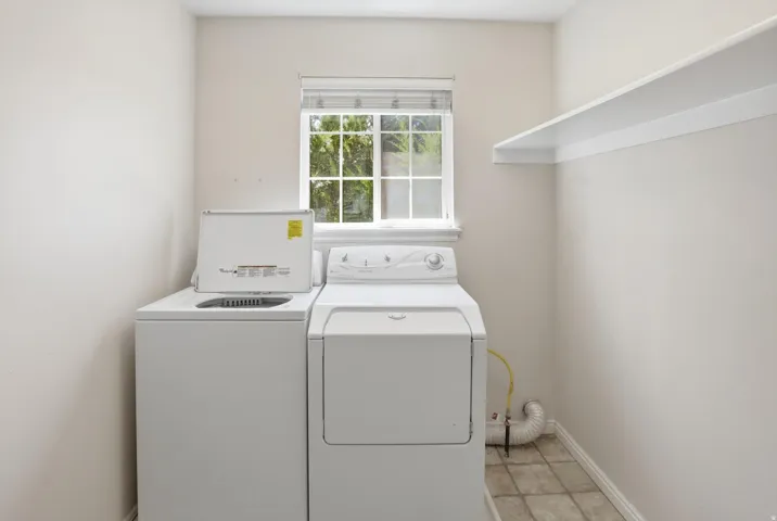 Dedicated utility space featuring a washer and dryer, a window with natural light, tiled flooring, and an elevated storage shelf