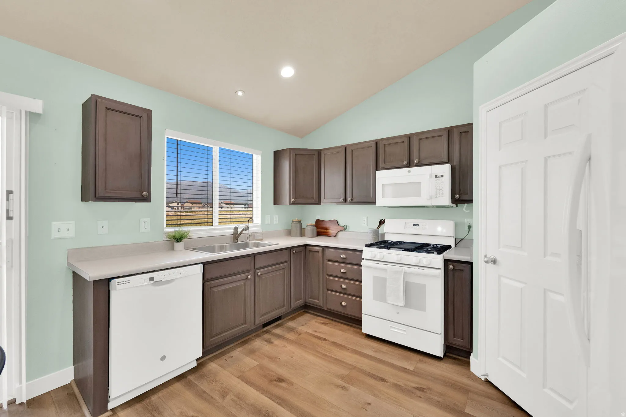 Kitchen featuring wood-finish flooring, white appliances, dark cabinetry, light countertops, and a window above the sink