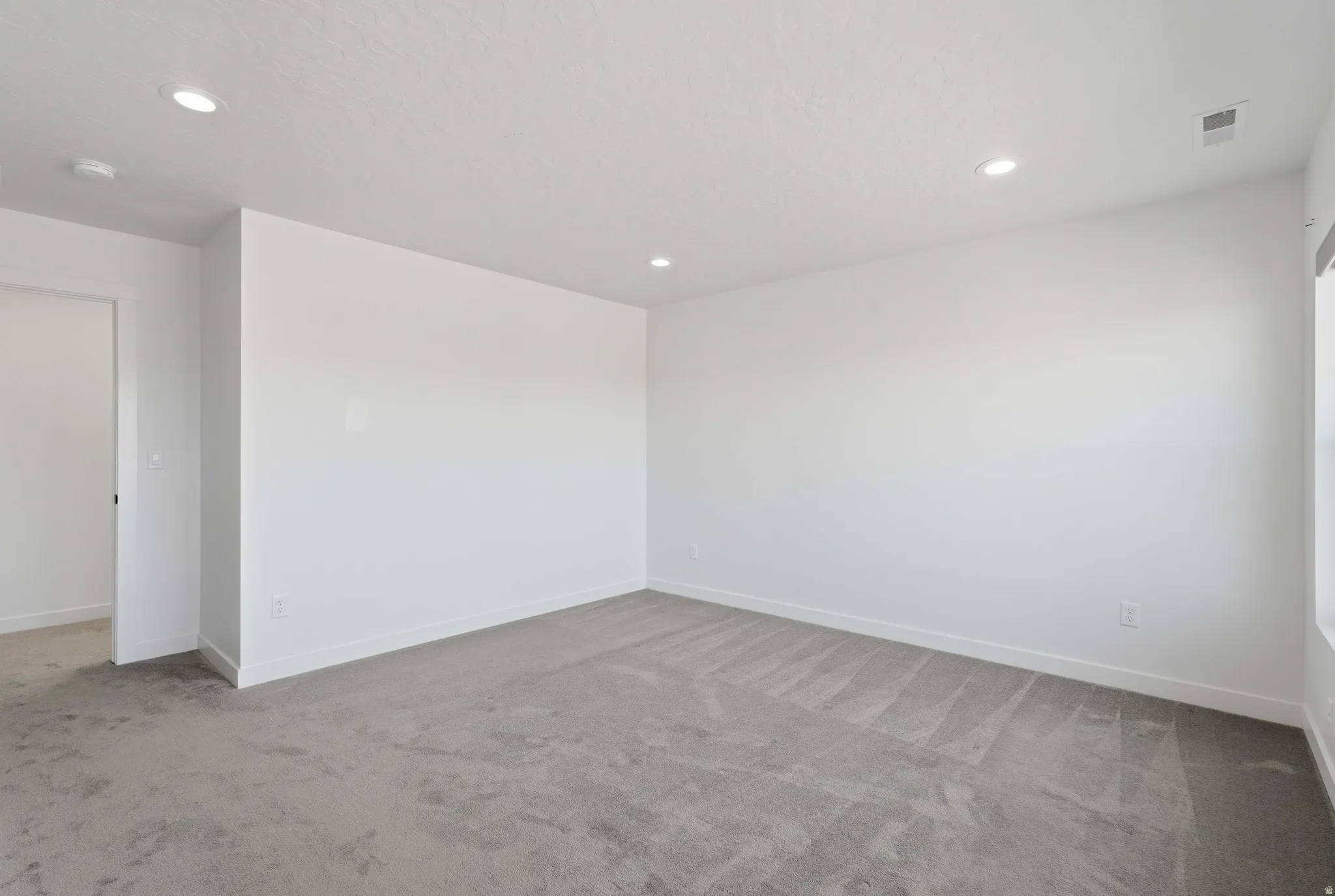 Spacious interior room featuring light gray carpeting, white walls, recessed lighting, and white baseboards