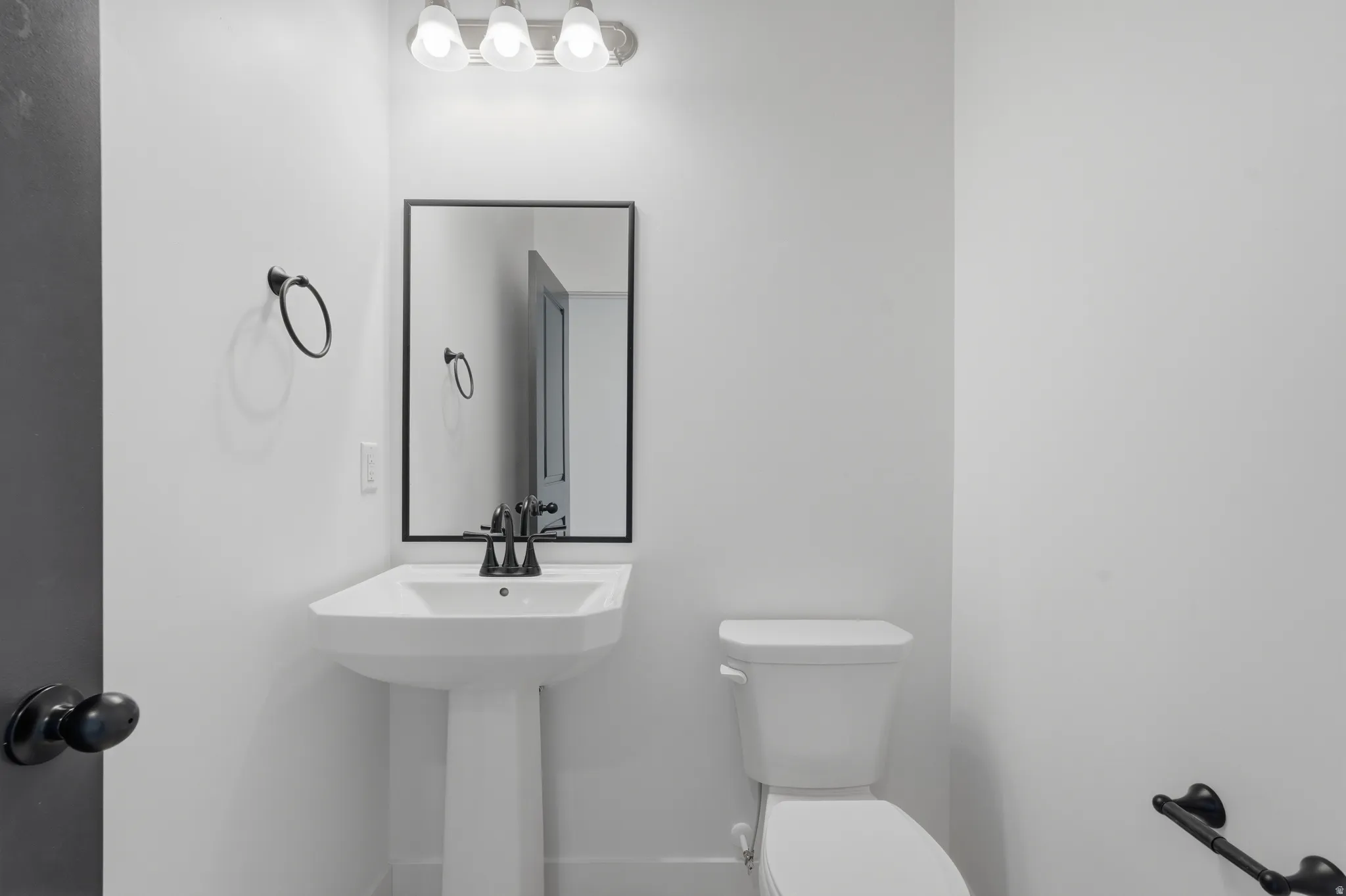 Pedestal sink with matte black faucet and coordinating mirror, complemented by a white toilet and modern light fixture