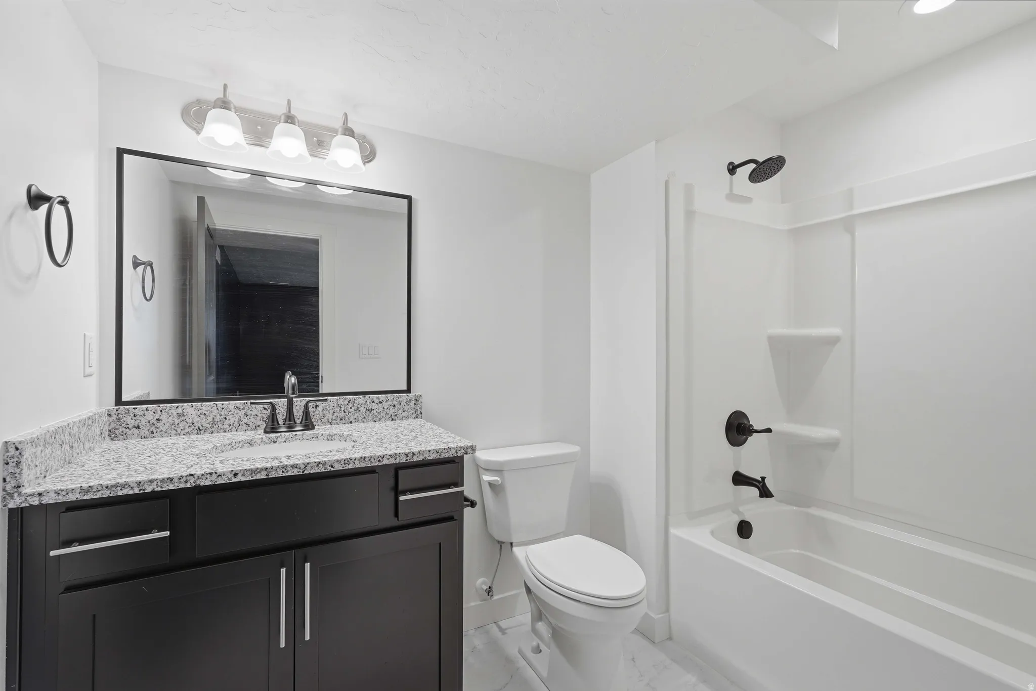 Full bathroom featuring a dark wood-finish vanity with a light speckled countertop, matte black fixtures, a framed mirror, and a full tub with shower