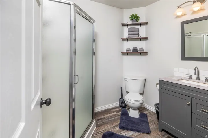Frosted glass shower enclosure, wood-finish flooring, dark gray vanity with granite-style countertop, subway tile backsplash, and corner-mounted floating shelves