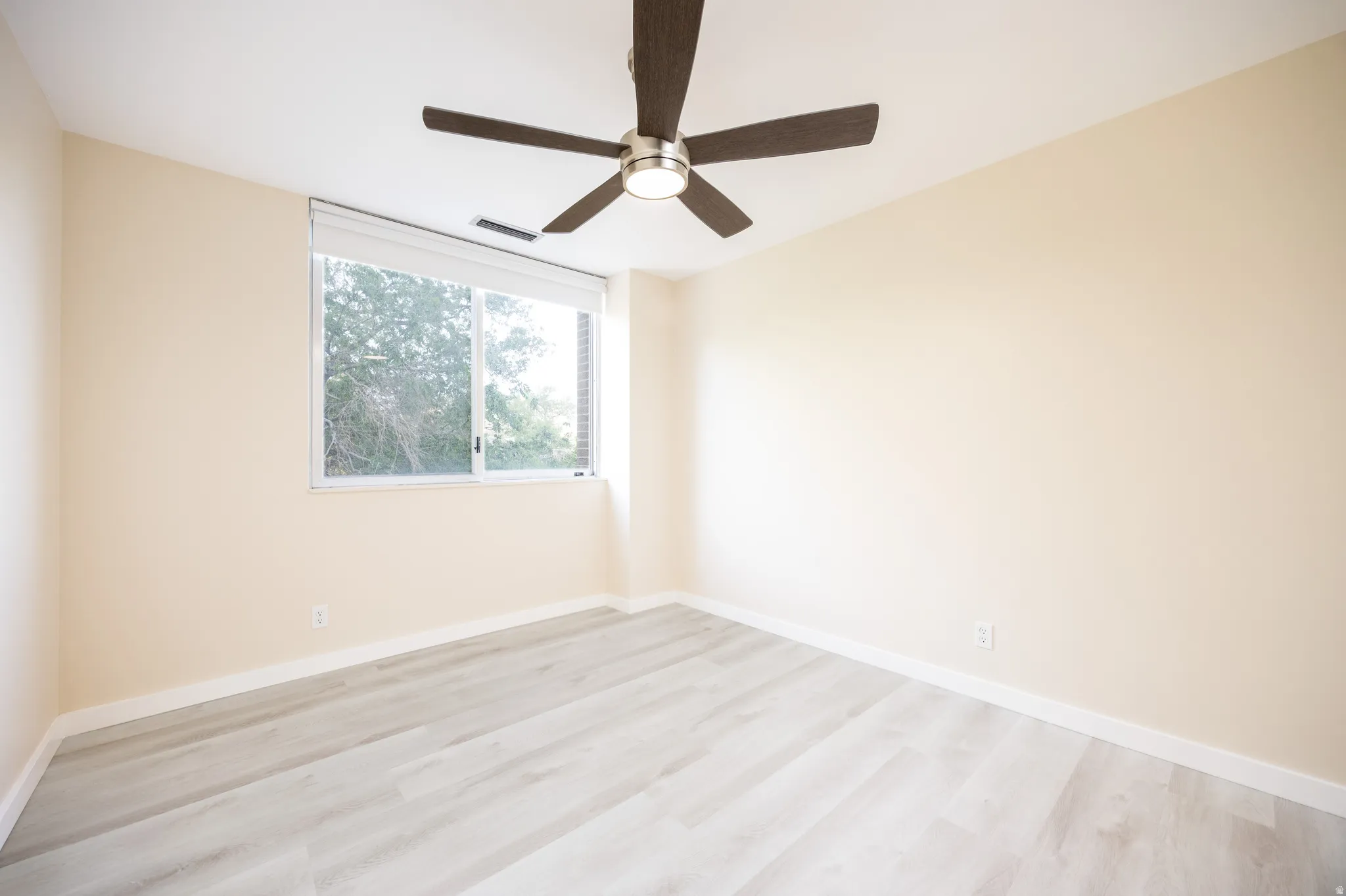 Bright room featuring a ceiling fan with integrated lighting, a large window with roller shades, light wood-finish flooring, and neutral wall paint