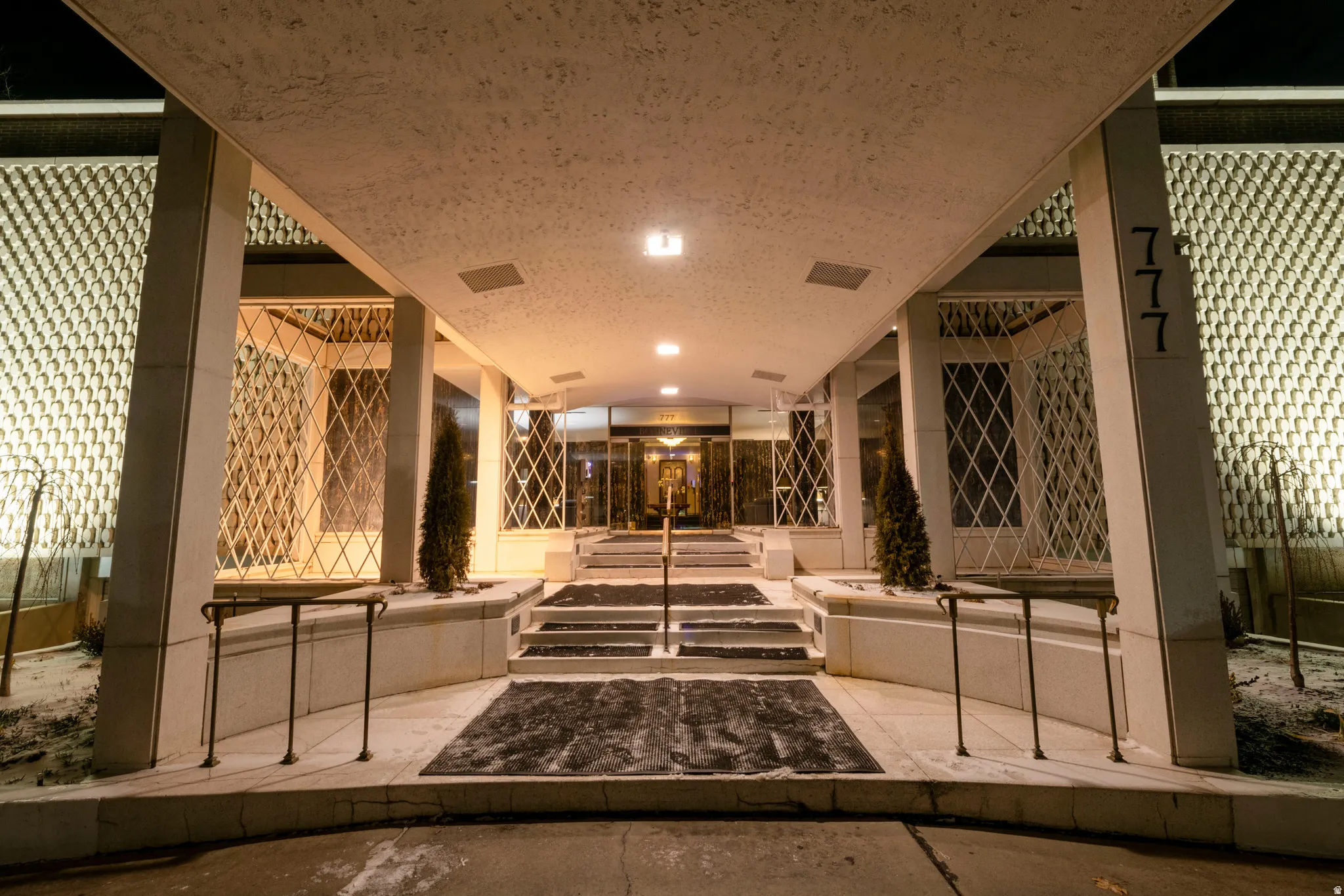 Covered portico entrance featuring recessed lighting, a multi-tiered concrete staircase, wrought iron railings, and decorative lattice panels