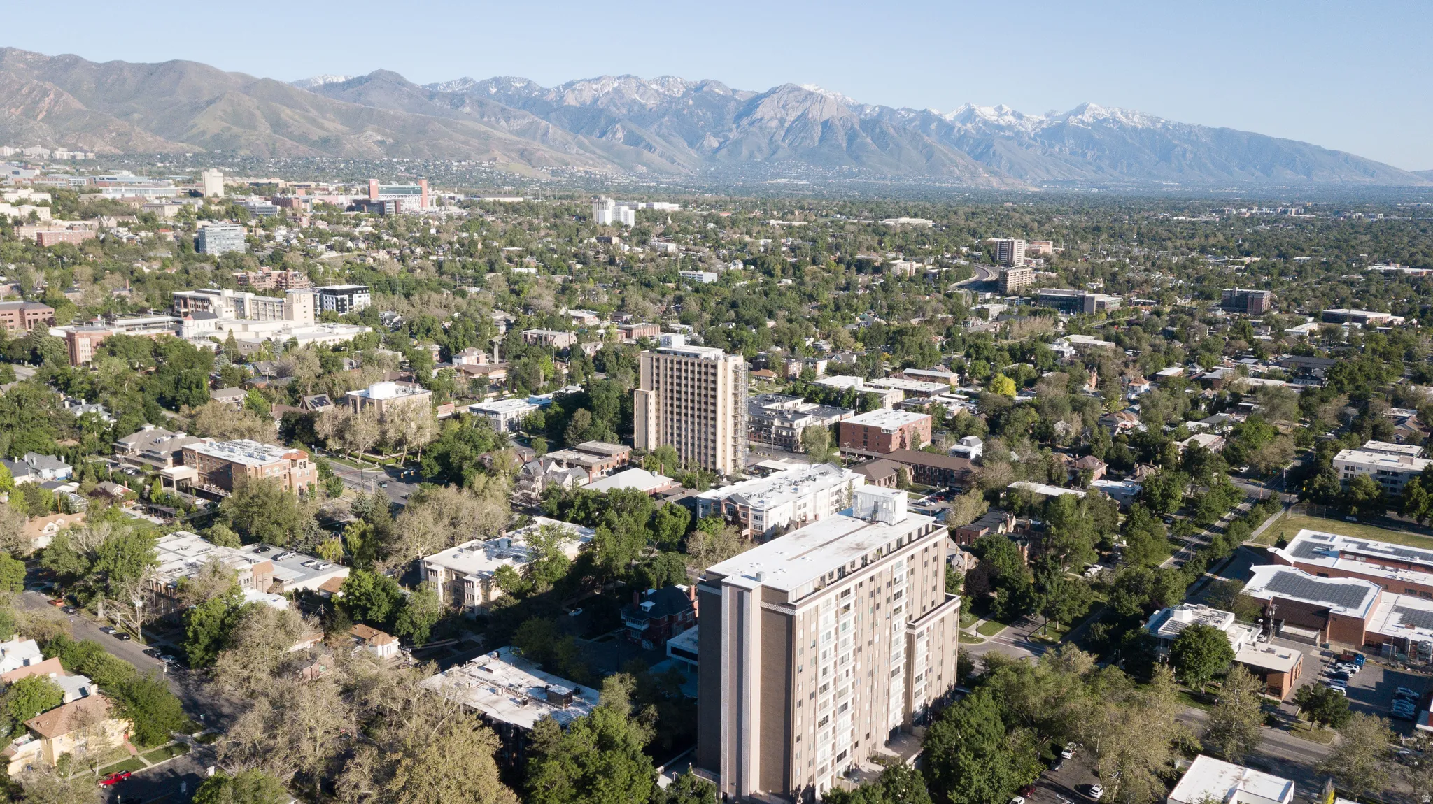 Expansive cityscape featuring a prominent multi-story building, abundant mature tree canopy, and distant snow-capped mountains