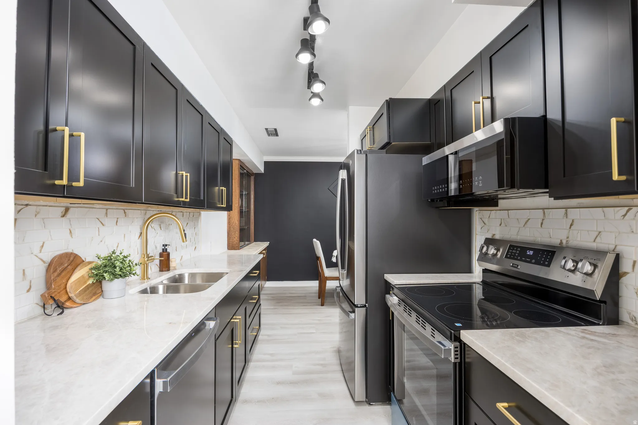 Kitchen featuring black cabinetry with gold-tone hardware, light-toned countertops, a stainless steel double-basin sink with a gold-tone faucet, a white and brown patterned backsplash, and light wood-finish flooring