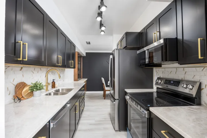 Kitchen featuring black cabinetry with gold-tone hardware, light-toned countertops, a stainless steel double-basin sink with a gold-tone faucet, a white and brown patterned backsplash, and light wood-finish flooring