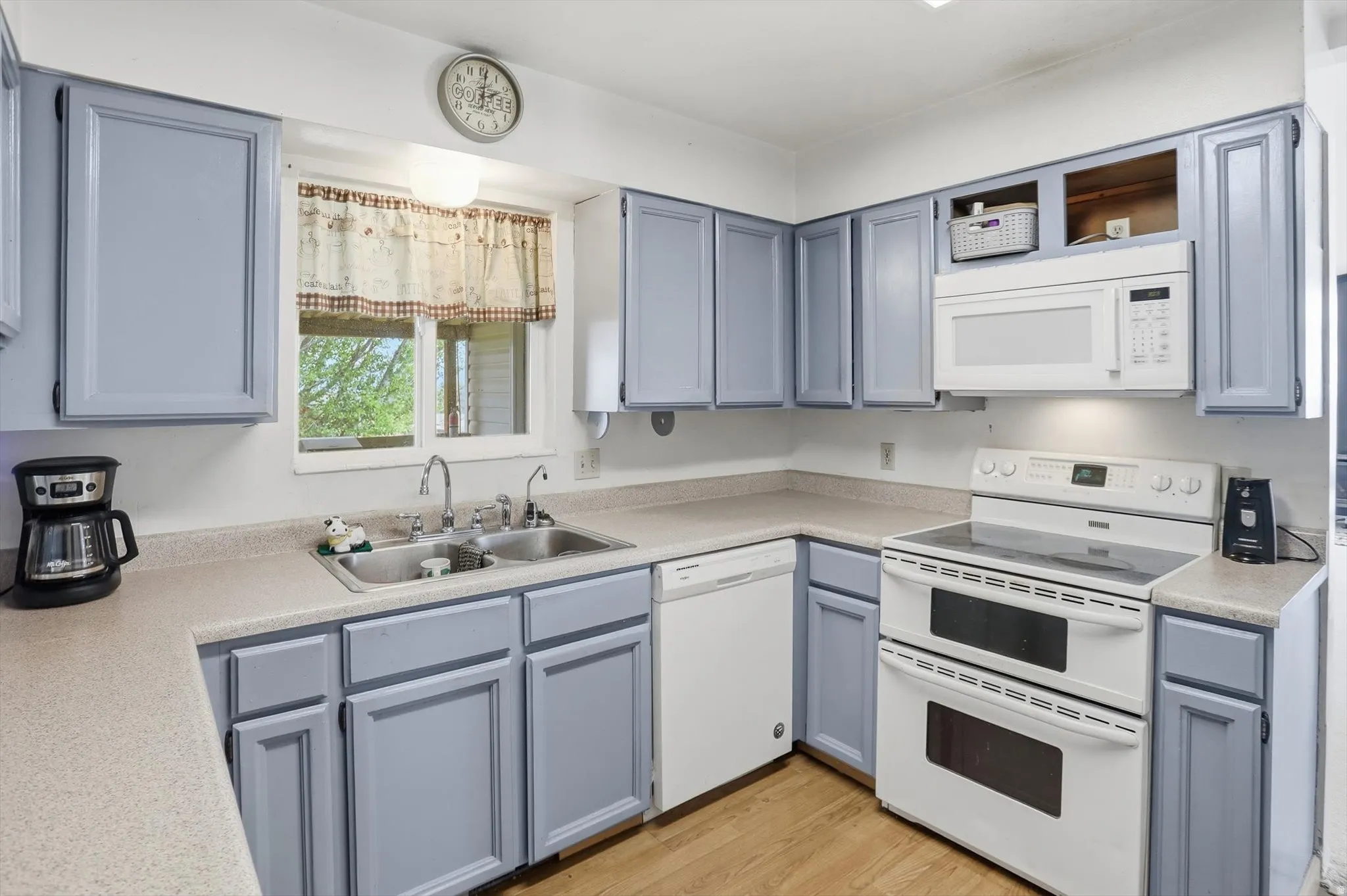 Kitchen featuring painted cabinetry, light-toned countertops, a double basin stainless steel sink, wood-finish flooring, and white appliances including a built-in microwave and dual-oven range