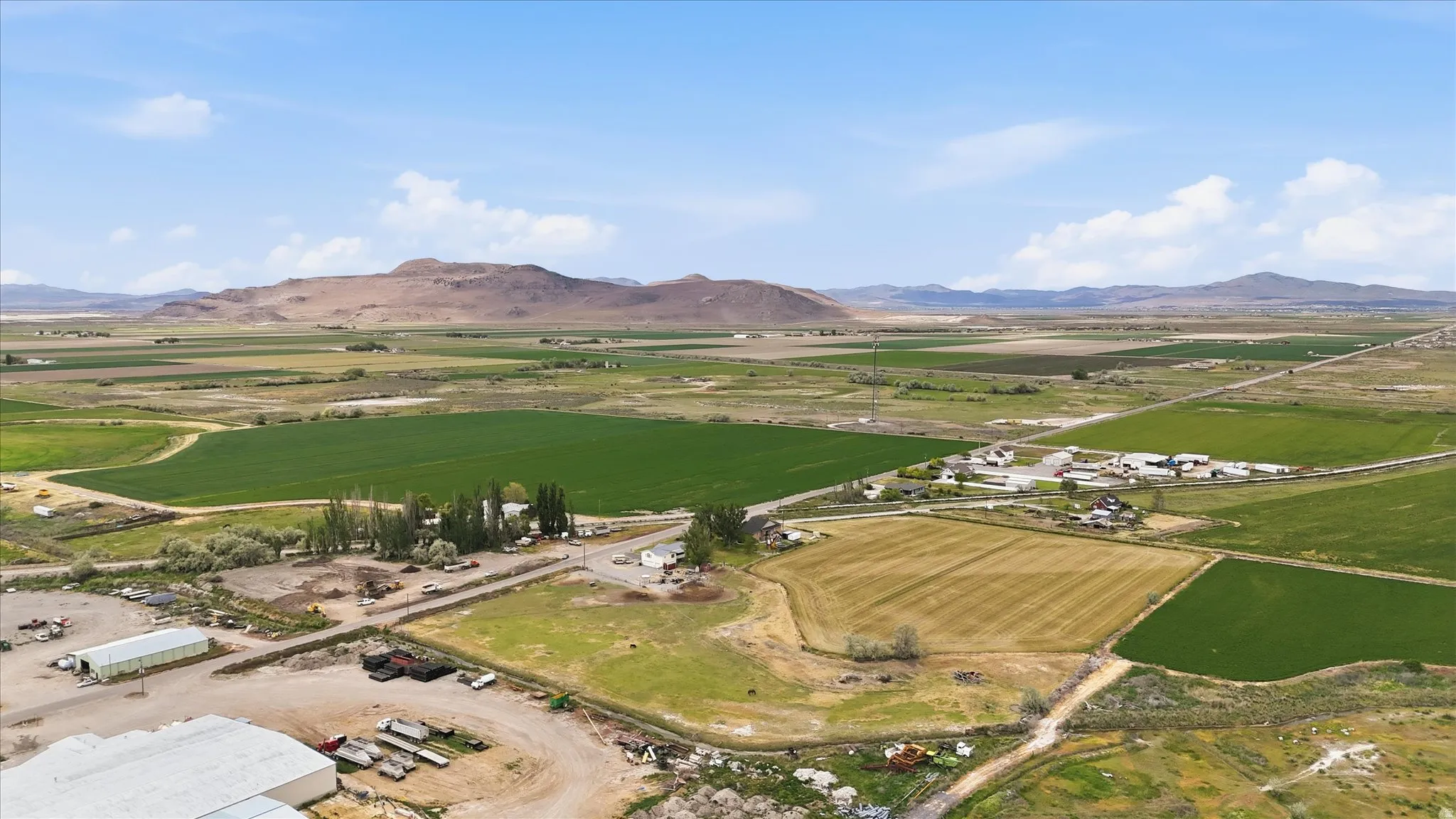 Expansive agricultural land featuring cultivated fields, scattered buildings, and distant mountains under a blue sky with clouds