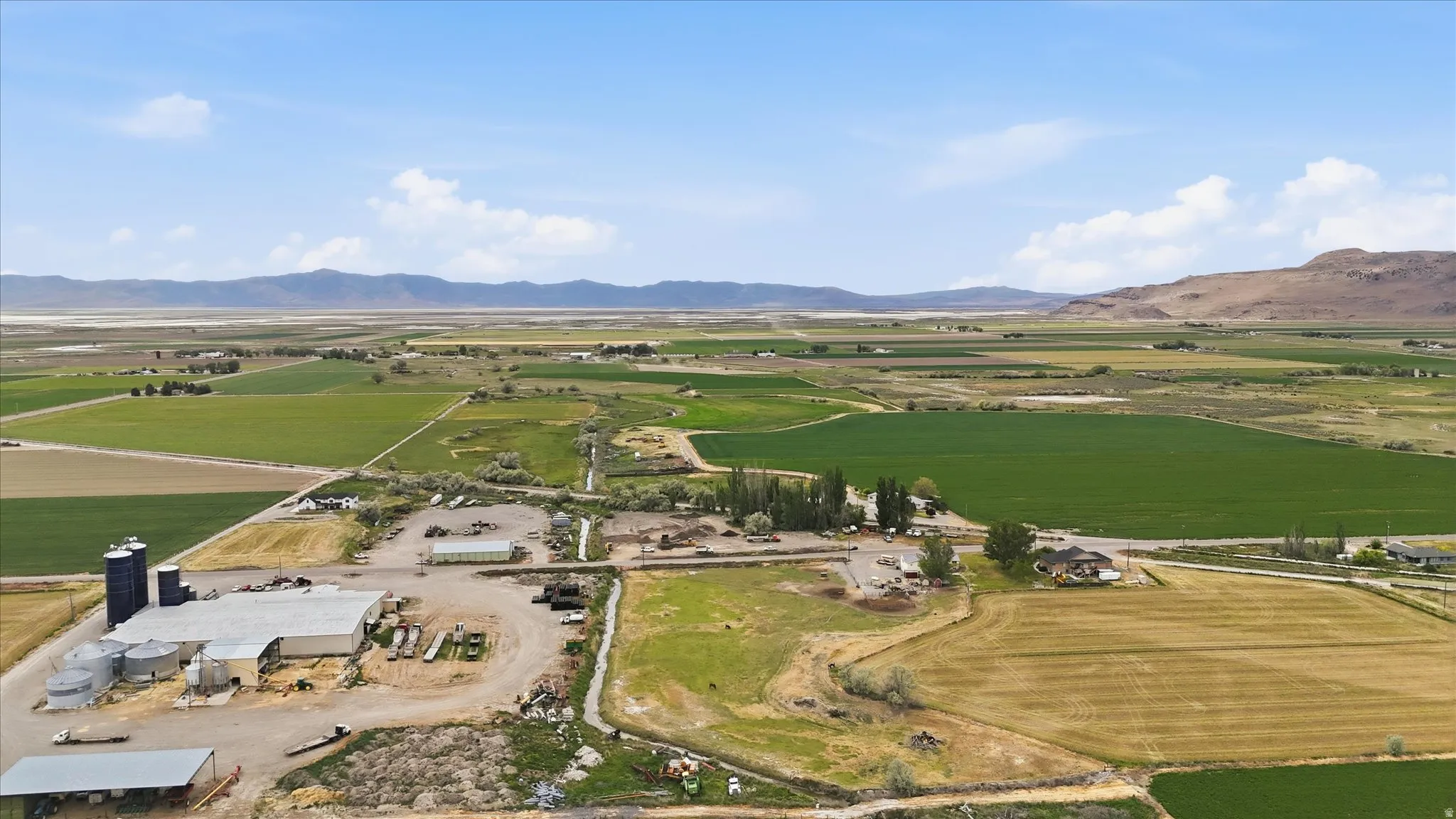 Expansive agricultural landscape featuring multiple cultivated fields, a large industrial building with several silos, and a distant mountain range under a clear sky