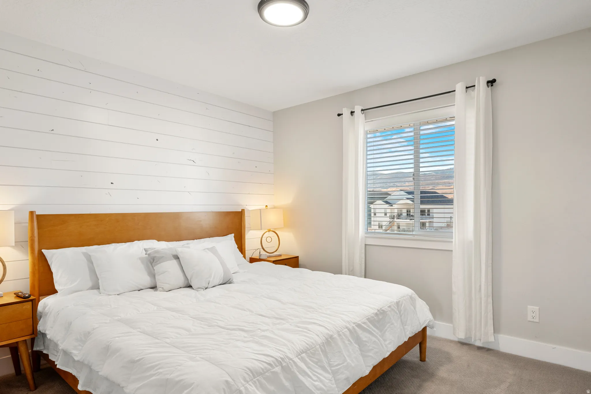 Bedroom featuring a prominent white shiplap accent wall, light-toned carpeting, and a window with white blinds and sheer curtains