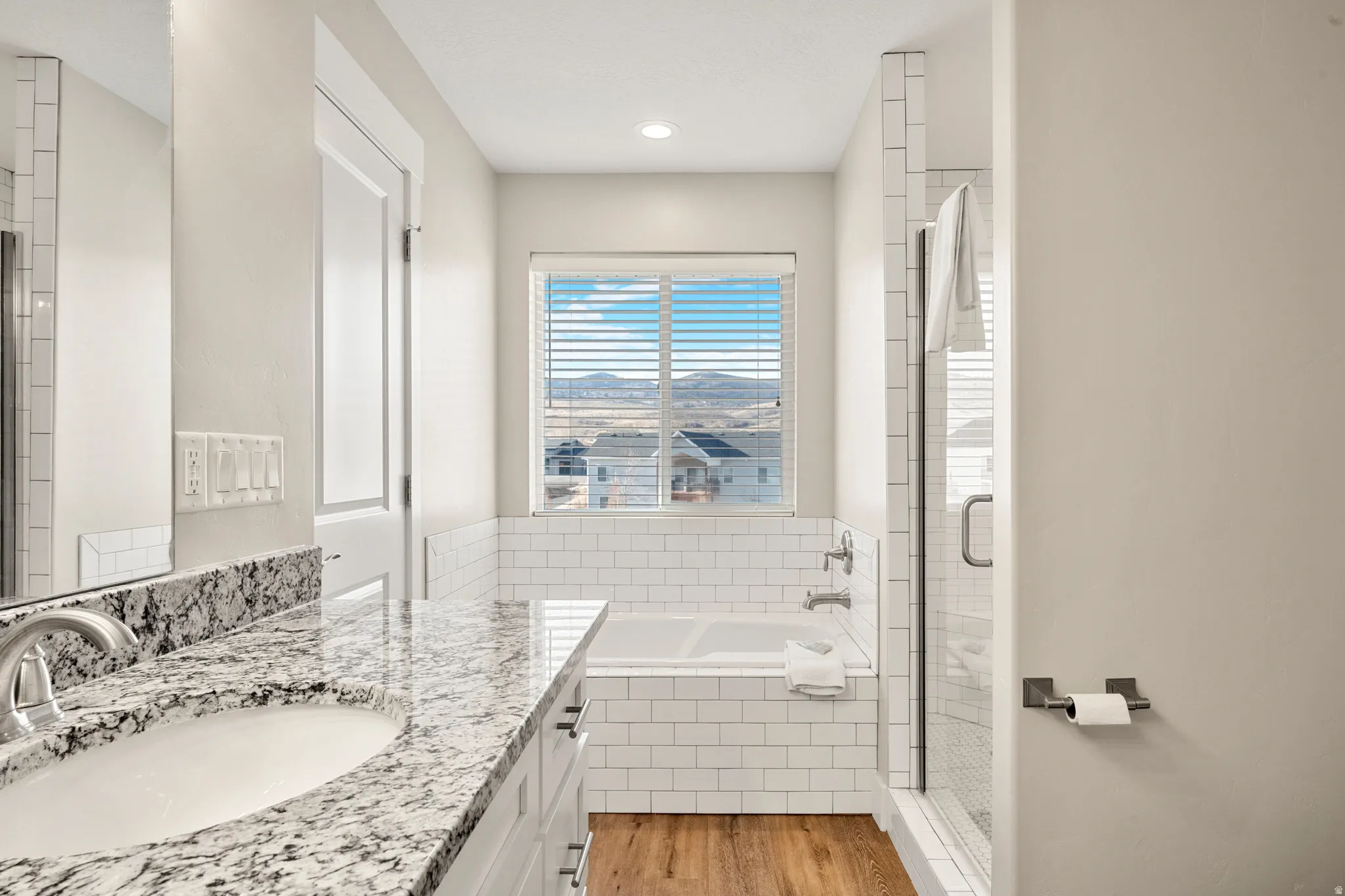 Modern bathroom featuring a double vanity with granite countertop and undermount sinks, wood-finish flooring, subway tile accents, a built-in tub, and a glass-enclosed shower