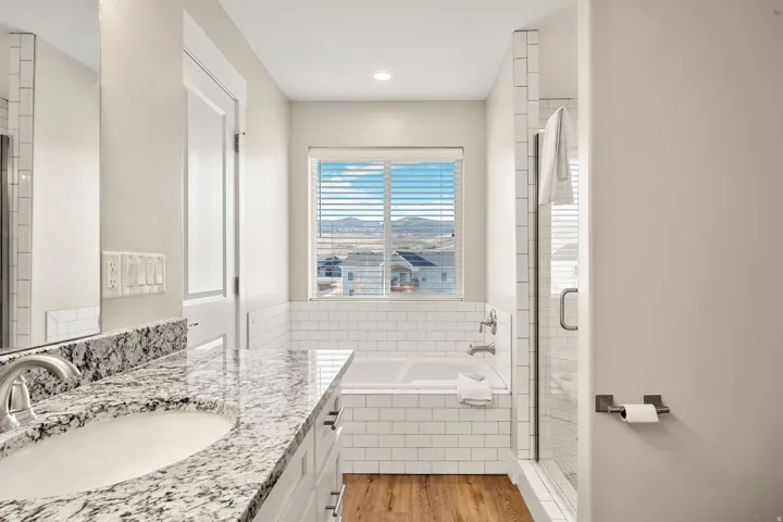 Modern bathroom featuring a double vanity with granite countertop and undermount sinks, wood-finish flooring, subway tile accents, a built-in tub, and a glass-enclosed shower