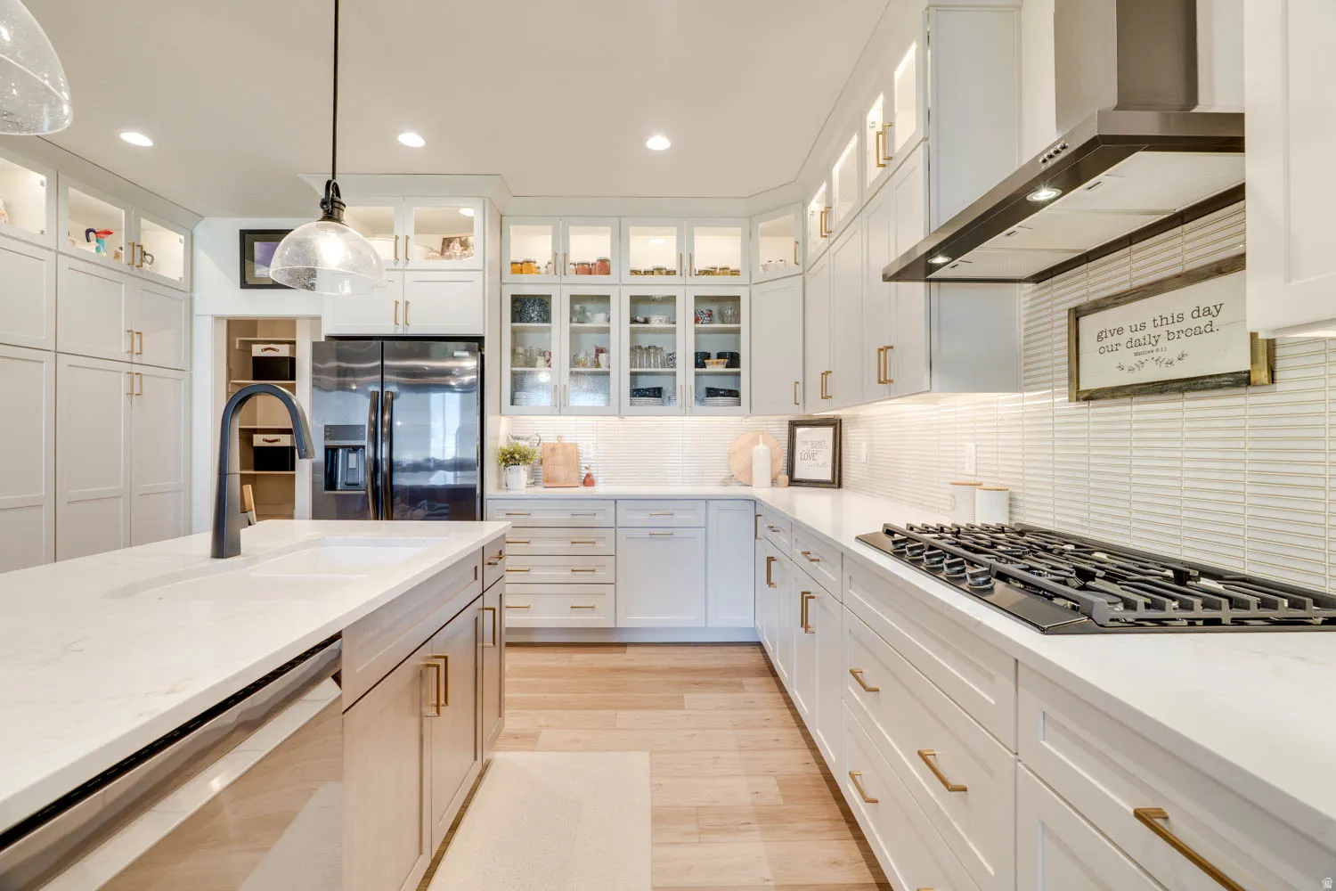 Open-concept kitchen featuring an expansive island with an undermount sink and matte black faucet, light wood-finish flooring, white cabinetry with gold-tone hardware, a five-burner gas cooktop, and a matching range hood