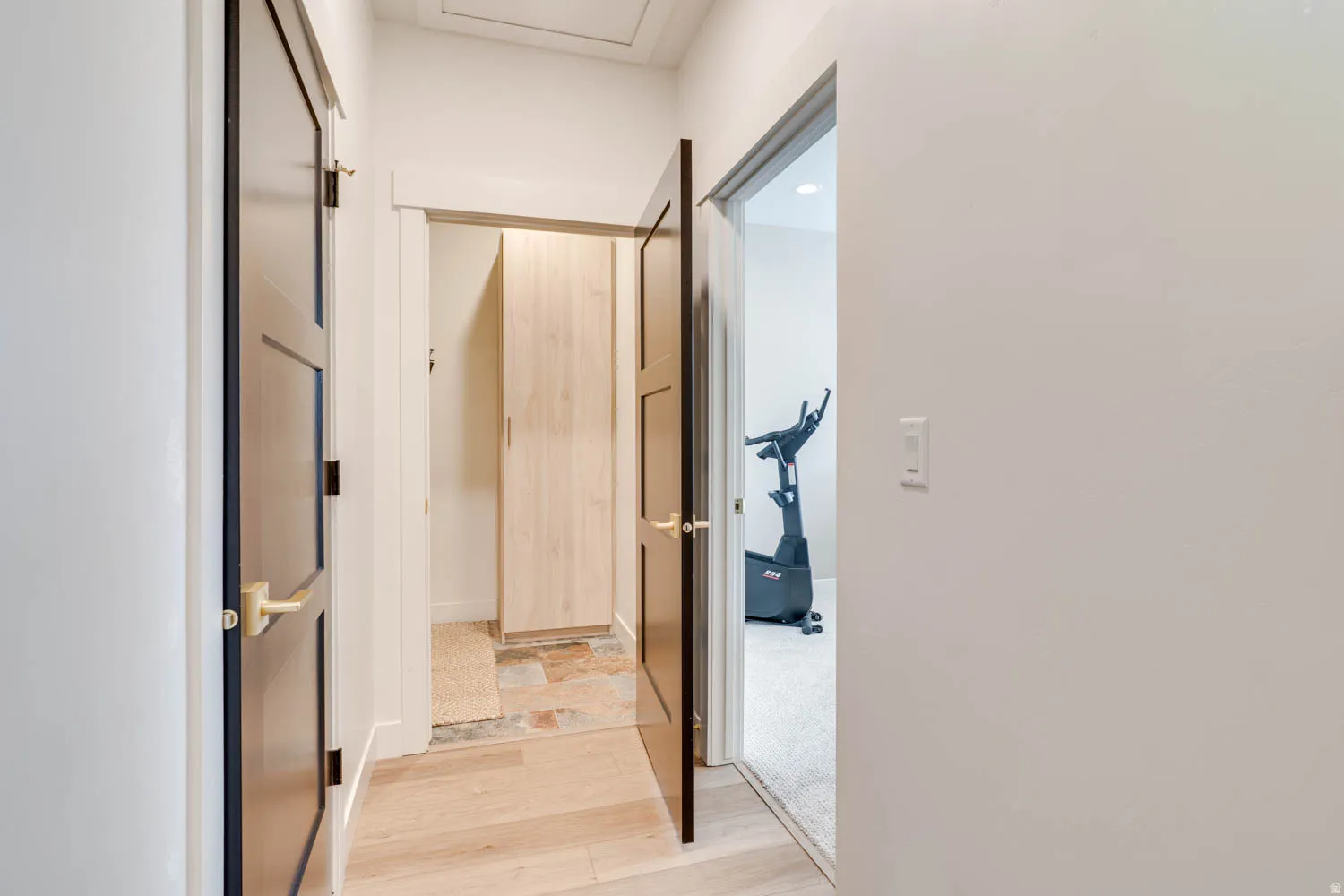 Hallway featuring light wood-finish flooring, white walls, and dark paneled doors with brass hardware