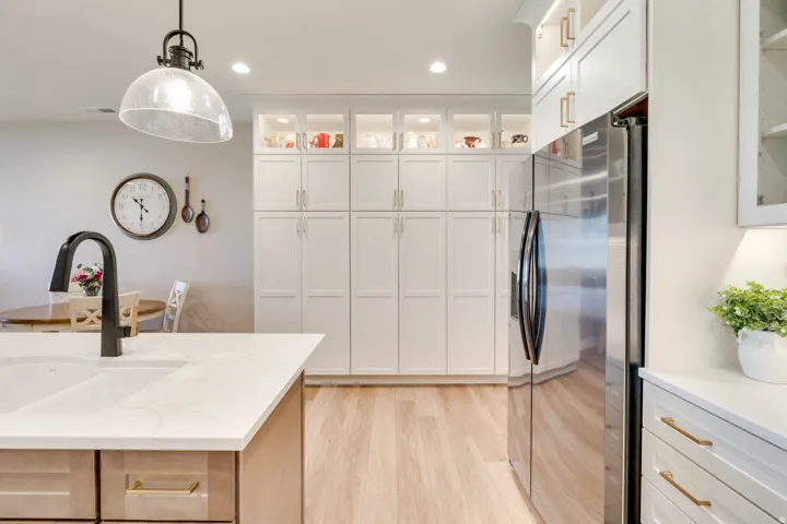 Contemporary kitchen featuring a large island with an integrated sink, a matte black gooseneck faucet, light wood-finish flooring, white shaker-style cabinetry with gold hardware, and a stainless steel refrigerator