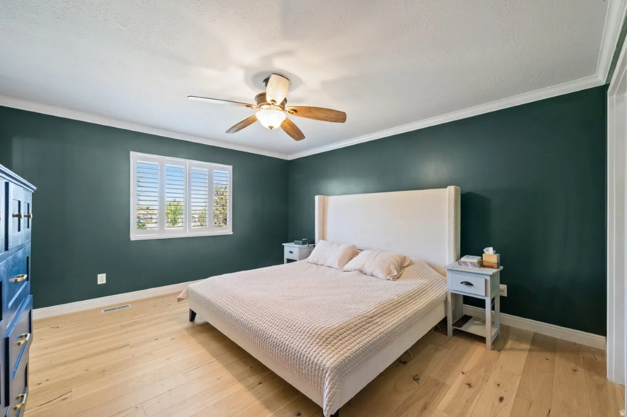 Bedroom featuring light wood-finish flooring, a ceiling fan with integrated lighting, and a window with plantation shutters