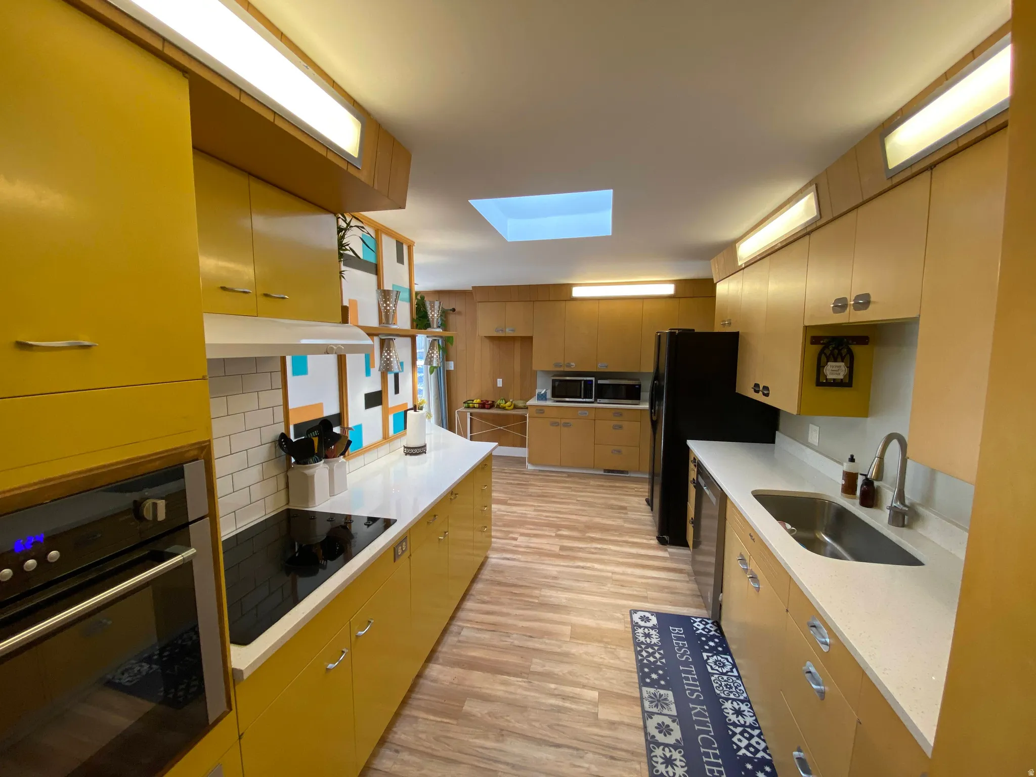 Kitchen featuring yellow and natural wood cabinetry, light countertops, wood-finish flooring, a built-in oven, and a black cooktop
