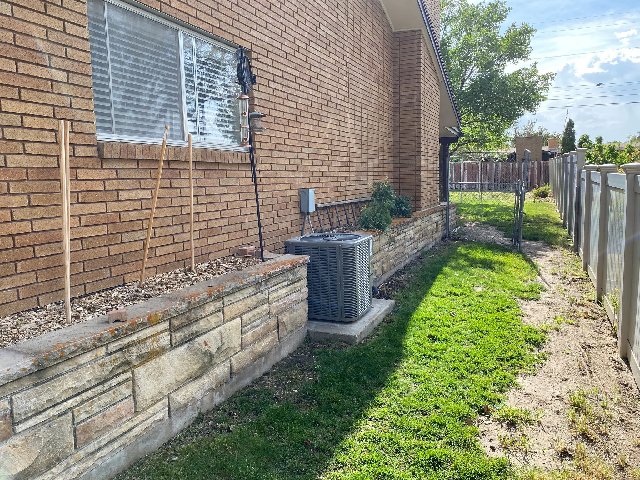 Brick exterior with a stone-clad planter, central air conditioning unit, and a chain-link gate providing yard access