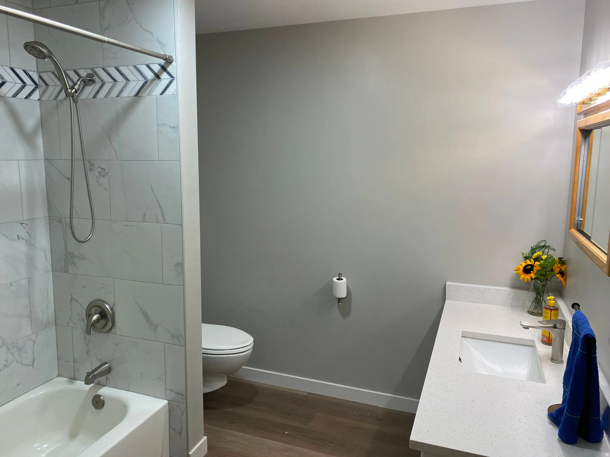 Bathroom featuring a white bathtub with a tile surround, a sleek vanity with an integrated sink, a light wood-finish mirror, and wood-finish flooring