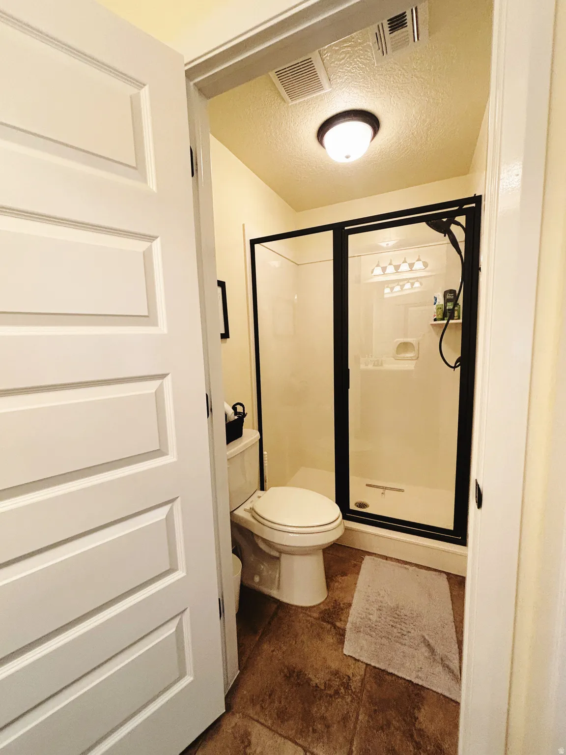 Bathroom featuring a stand-up shower with a dark-framed glass enclosure, white toilet, and textured flooring