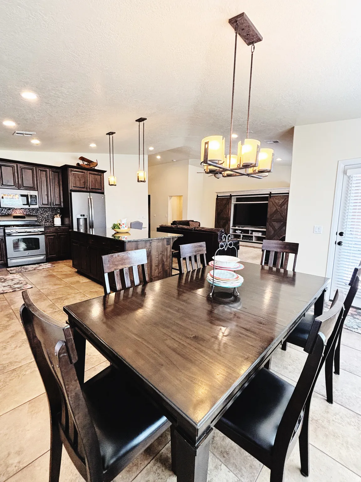 Open-concept living space with tile flooring throughout, featuring a kitchen with dark wood cabinetry and stainless steel appliances, a kitchen island with seating, and a dining area illuminated by a multi-light chandelier