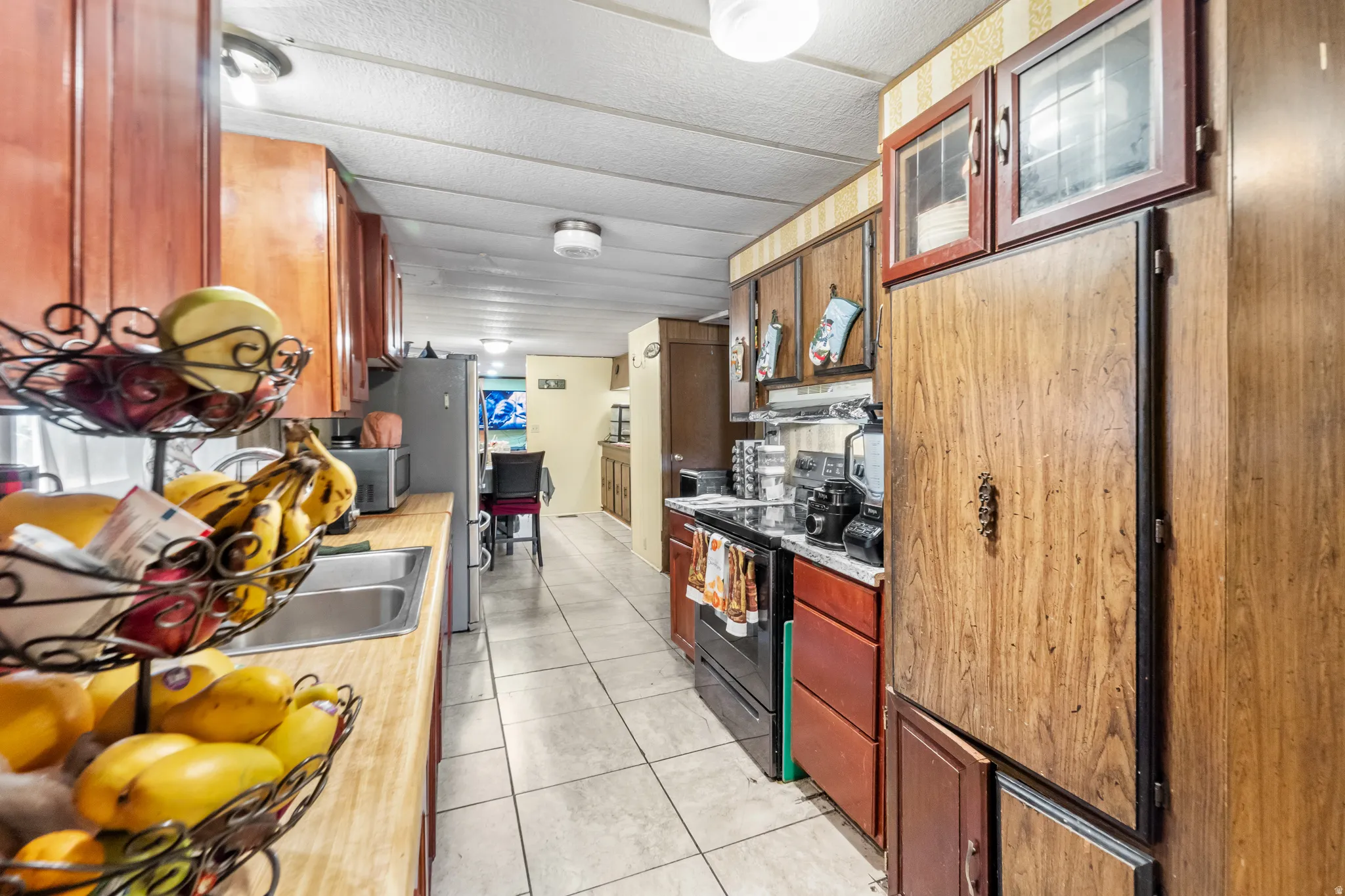 Galley kitchen featuring wood-finish cabinetry, a double basin stainless steel sink, a black electric range, and light-toned tile flooring