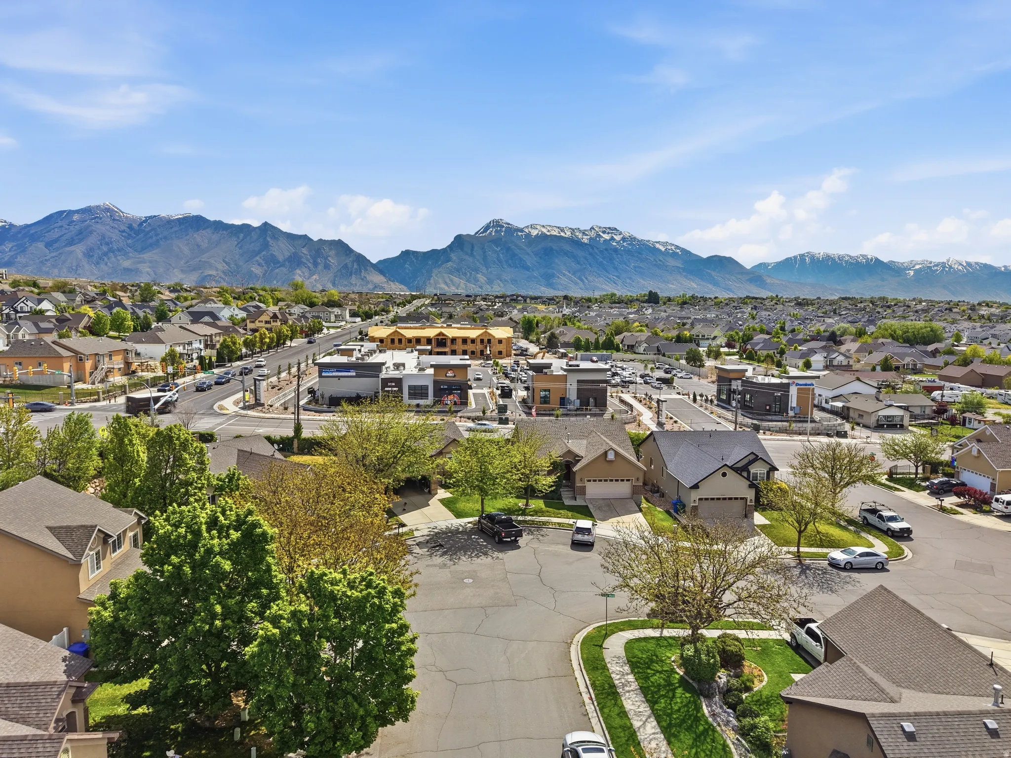 Expansive suburban landscape featuring a mountainous backdrop, residential properties with varied architectural styles, commercial buildings with modern facades, a network of paved roads, and mature trees throughout