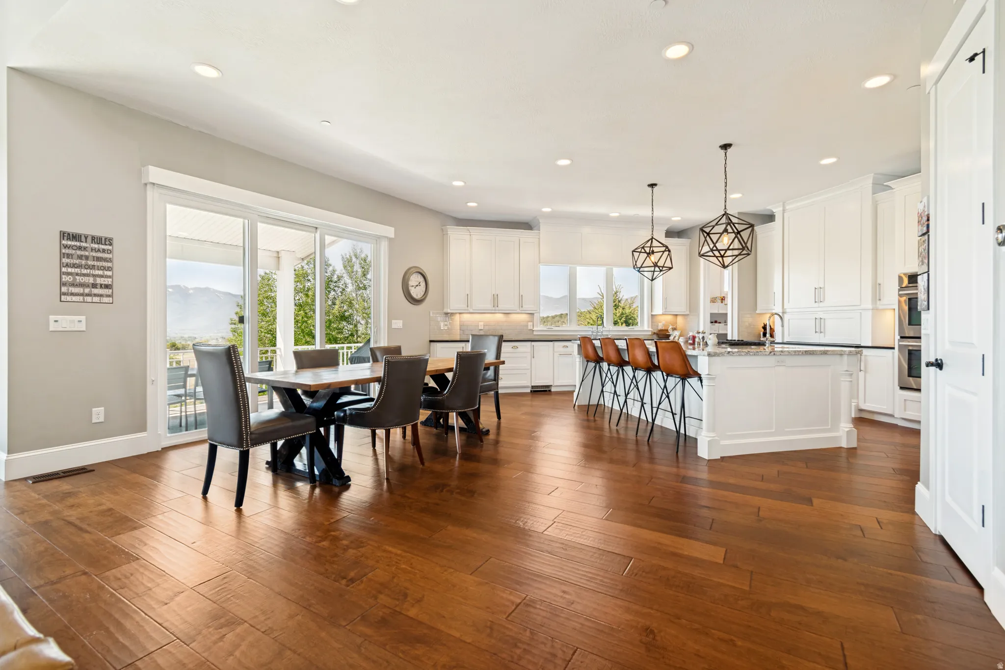 Open-concept living space featuring wood-finish flooring, white kitchen cabinetry with built-in appliances, a kitchen island with seating, pendant lighting fixtures, and sliding glass doors revealing a mountain backdrop