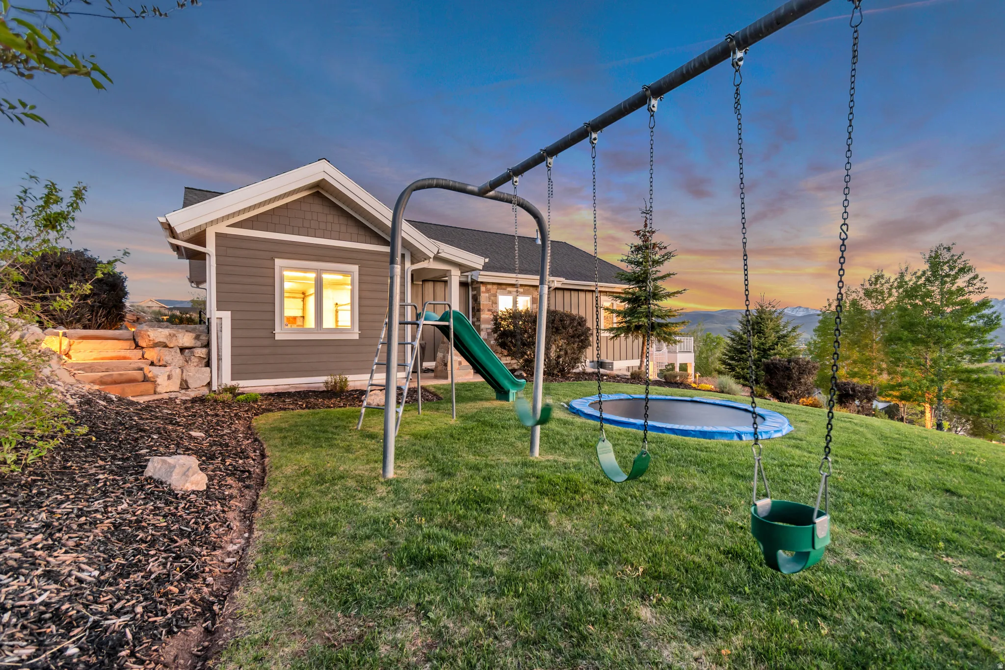 Expansive green lawn with a swing set and trampoline, backed by a wood-sided dwelling featuring a stone retaining wall and landscaped terraced steps