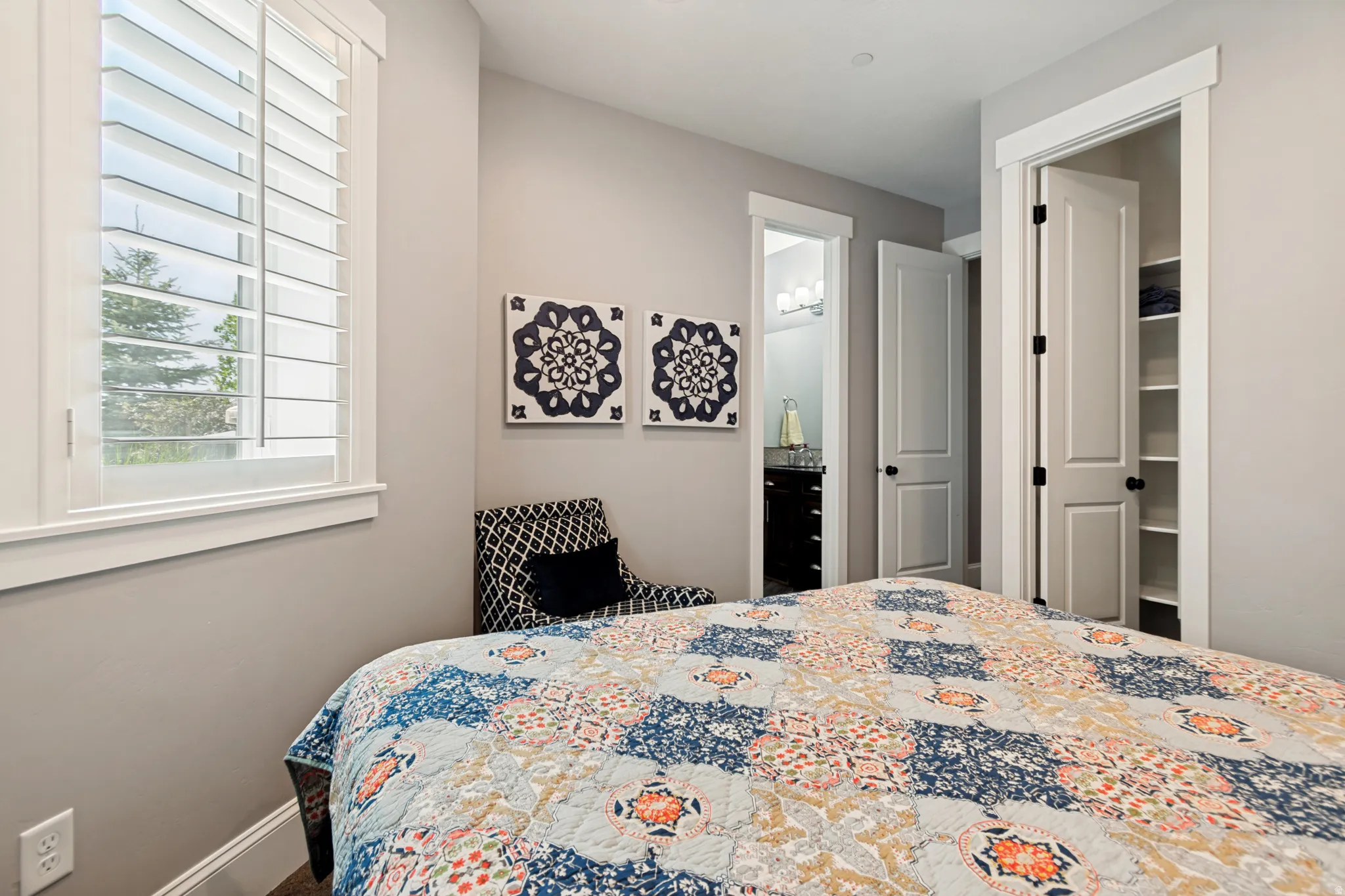 Interior room featuring neutral wall tones, white trim, a window with plantation shutters, a door leading to a bathroom with a vanity and lighting, and a closet with built-in shelving