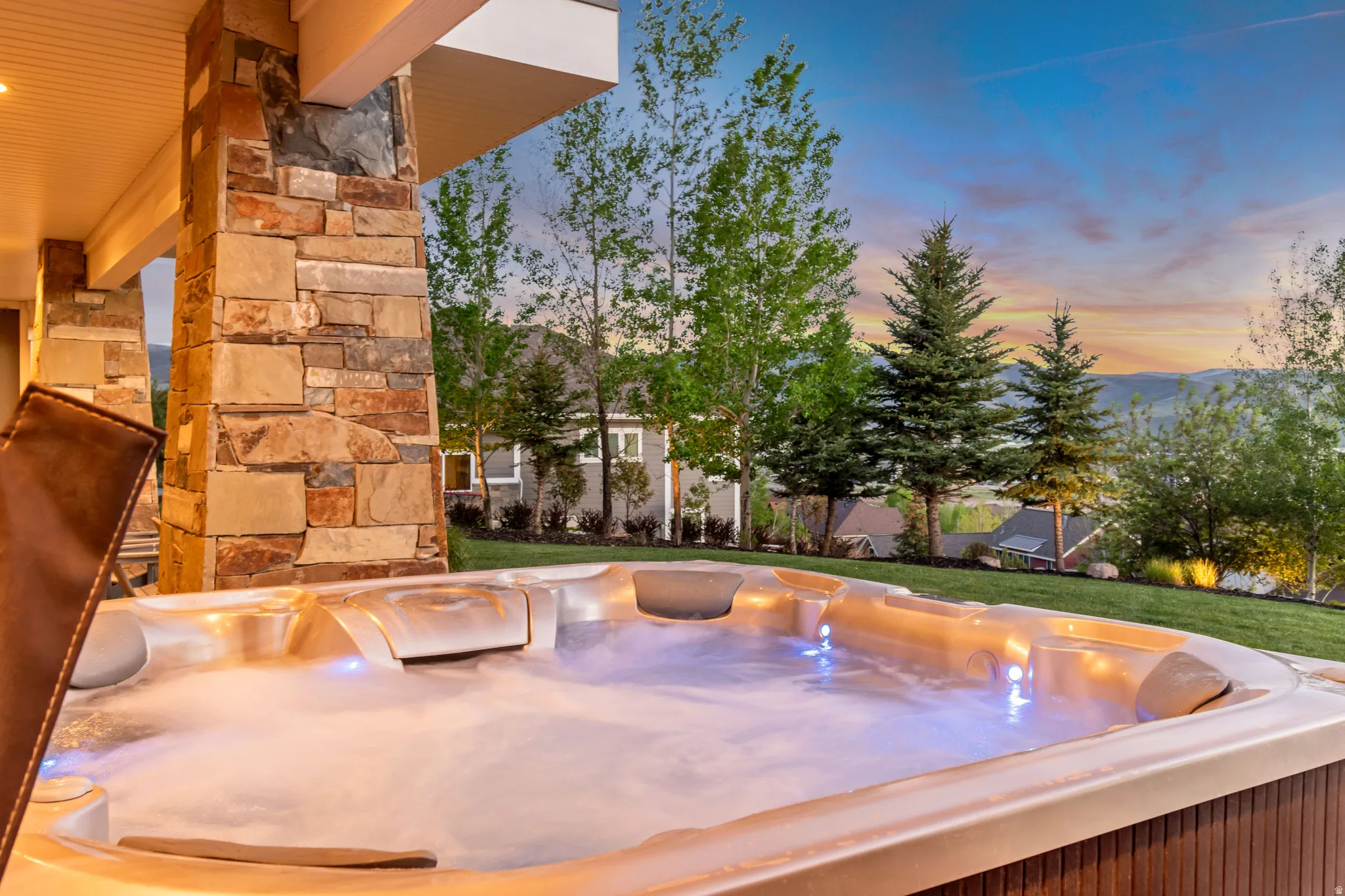 Outdoor hot tub with integrated lighting, a stone-clad column, an overhead patio structure, a manicured lawn, and distant mountain scenery