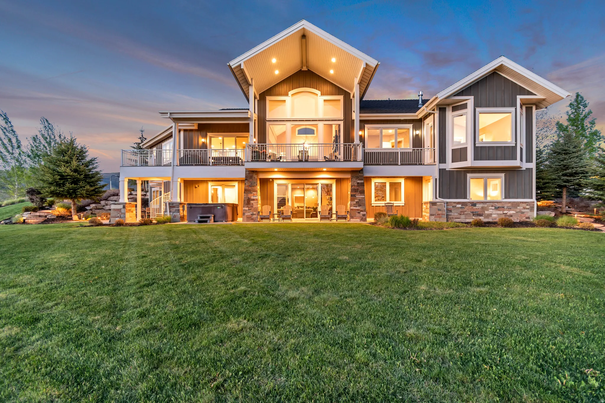 Contemporary multi-level residence featuring dark siding with stone accents, a prominent upper-level balcony, a lower-level patio, a spa, and extensive landscaping