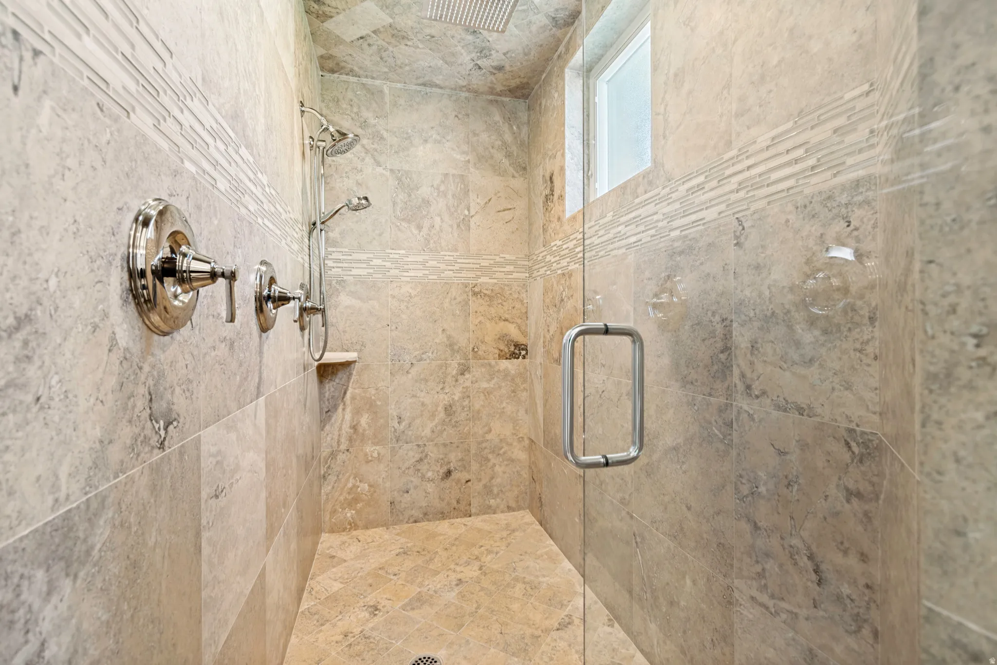 Shower with full-height stone-look tile, glass enclosure, polished chrome fixtures, integrated shelf, and a frosted window