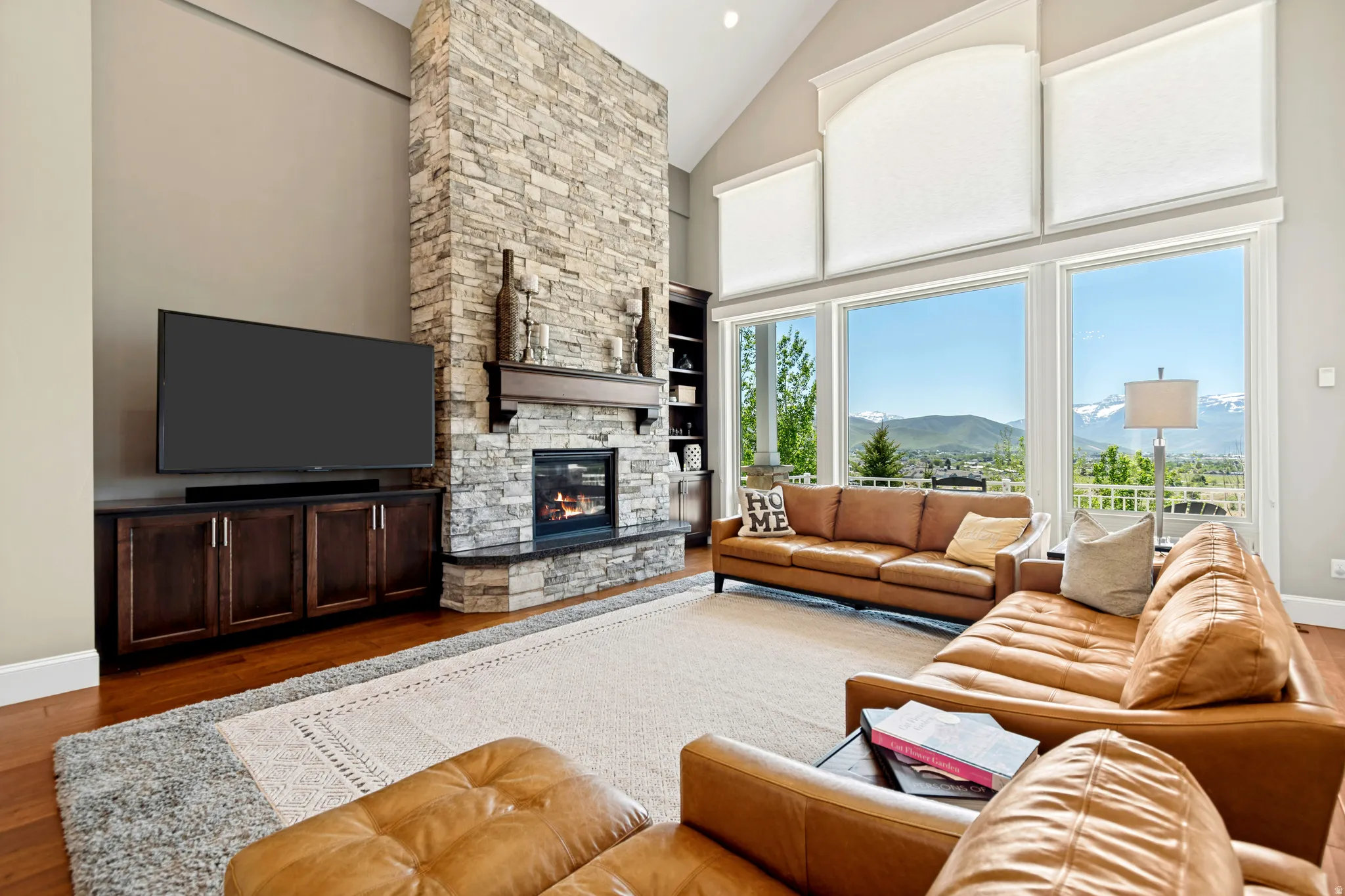 Vaulted great room featuring a floor-to-ceiling stone fireplace with a wood mantle and hearth, built-in media cabinetry, and high windows overlooking mountain scenery