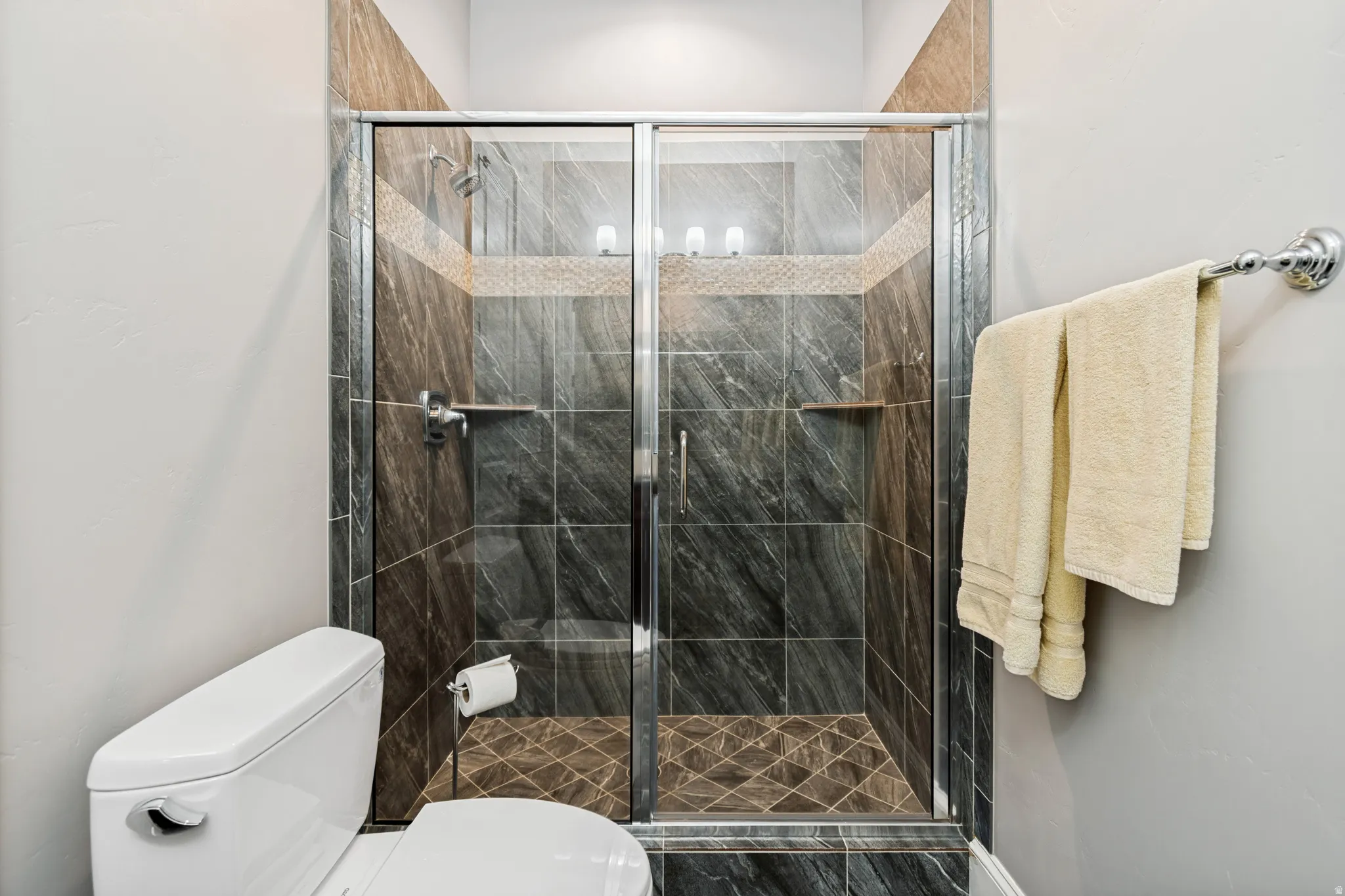 Glass-enclosed shower featuring dark stone-look tile, patterned floor tile, and a decorative tile accent band